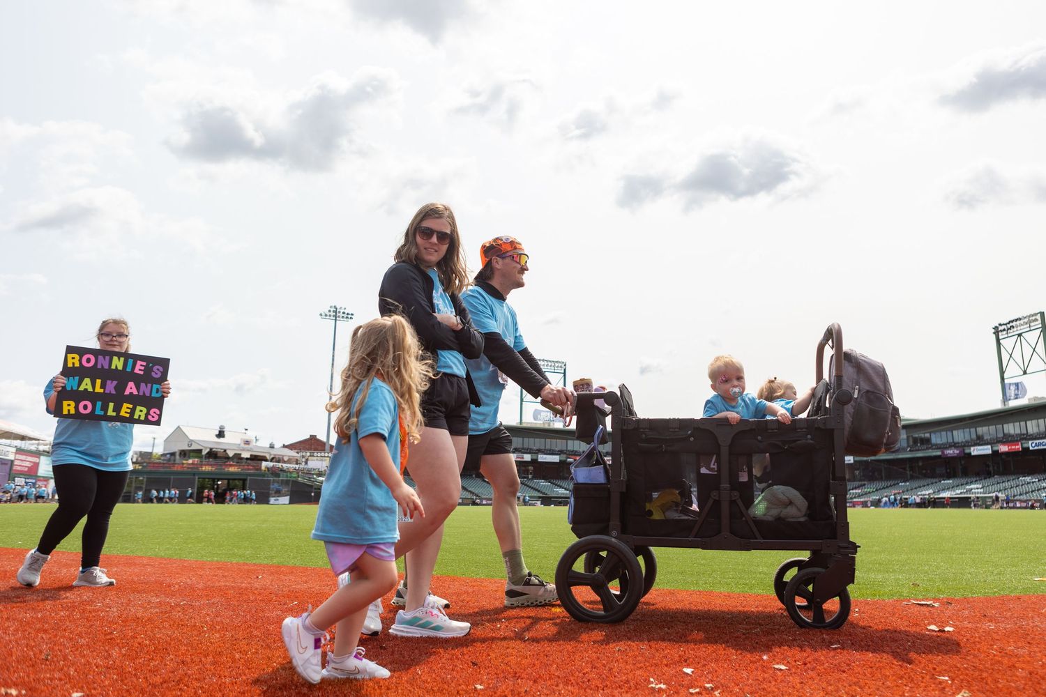 People navigate the baseball field with a large utility wagon during a stadium event, orange clay turf visible beneath them.