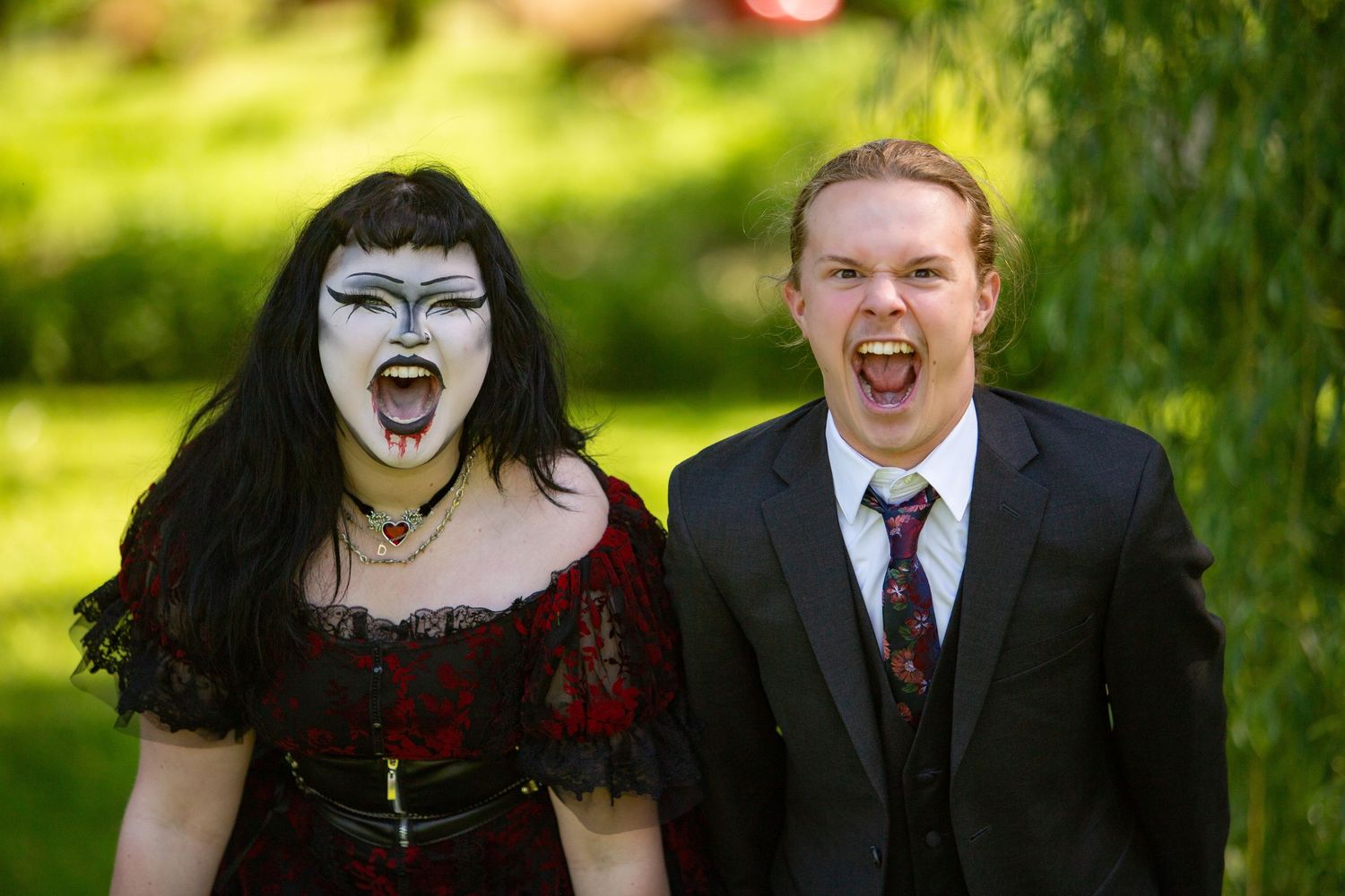 Two people in gothic and formal attire share an exuberant laughing moment outdoors in a sunlit garden setting.