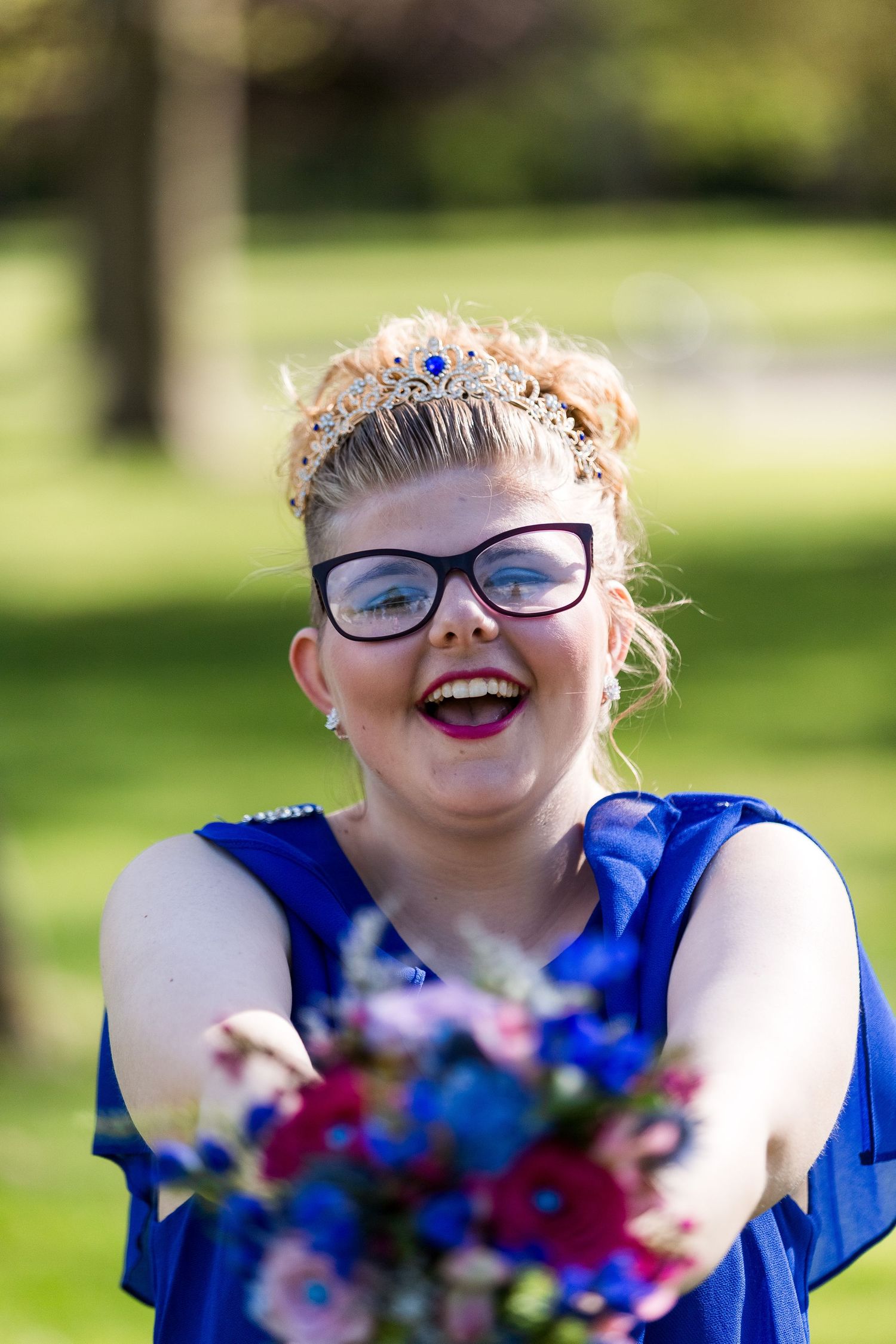 Person wearing glasses and blue dress holds out vibrant red and blue bouquet while smiling outdoors in a park setting.