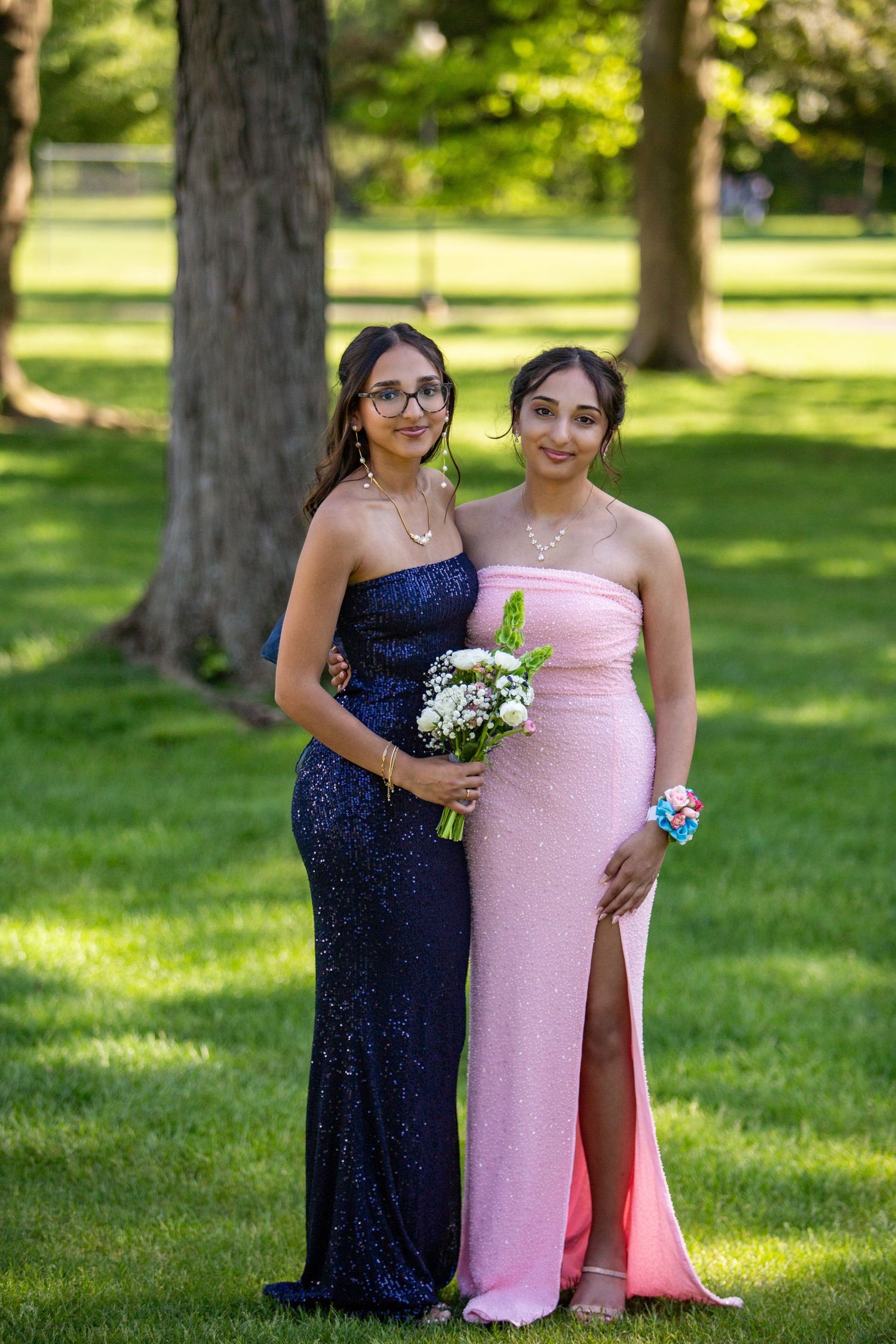 Two elegantly dressed figures pose at a formal event in a park, wearing a navy sequined gown and pink dress with floral corsages.