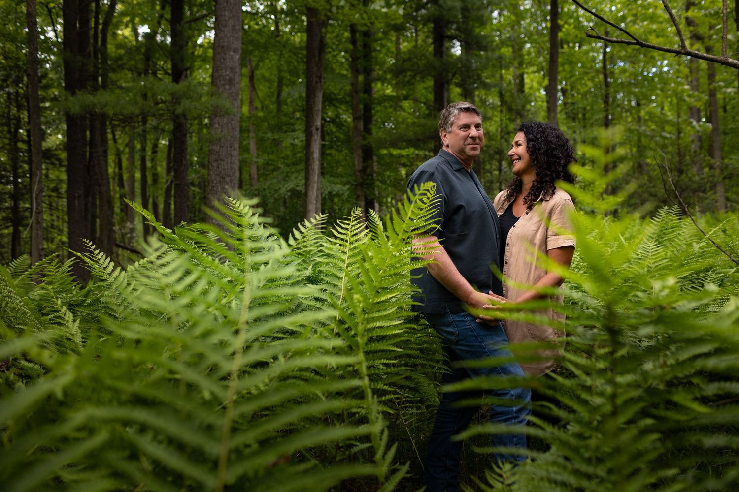 A romantic engagement photo session shows a couple surrounded by lush green ferns in a forest setting.