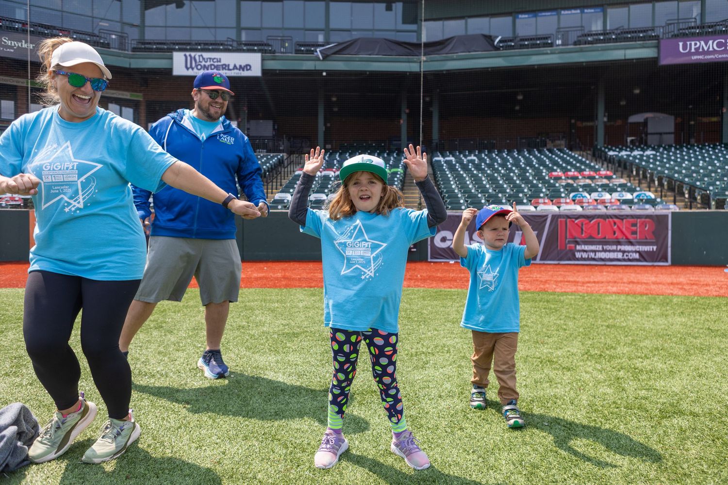 Families in matching blue shirts dancing together on baseball field during community event.