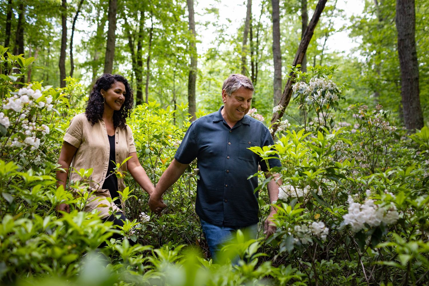 Two people hold hands while walking through a lush forest filled with blooming rhododendrons and green foliage.