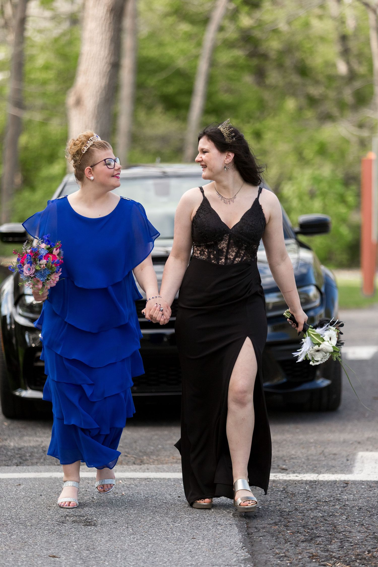 Two women in formal dresses, one in royal blue and one in black with a slit, walking together while holding hands.