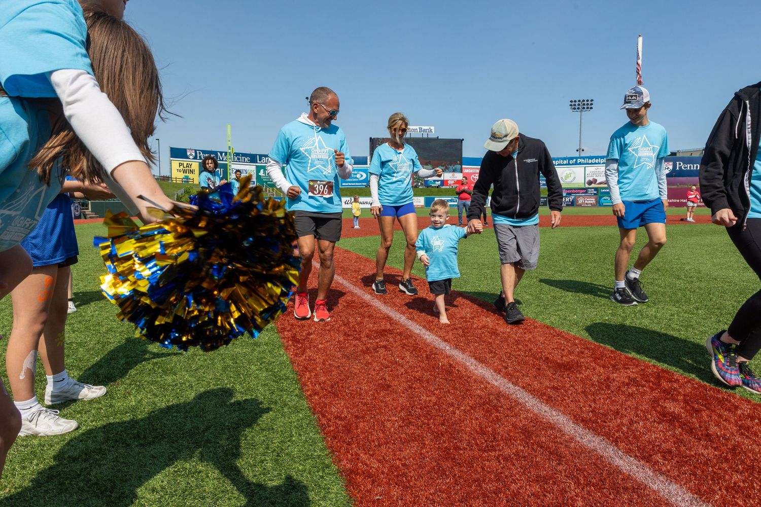 Participants in light blue shirts walk along a red track at a sports field while cheerleaders with pom-poms cheer.