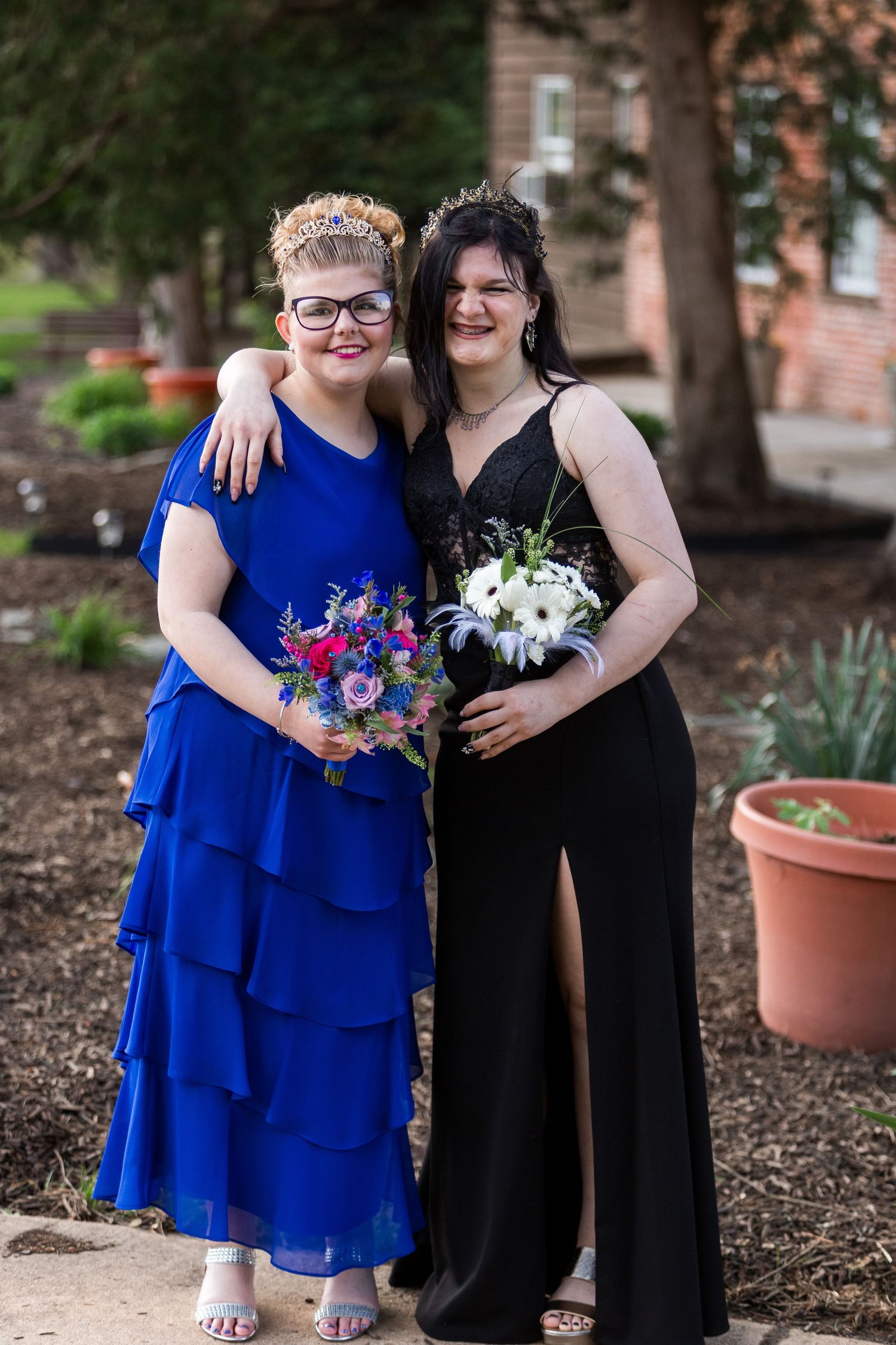 Two people in formal dresses with bouquets pose together at an outdoor event, one in royal blue and one in black.