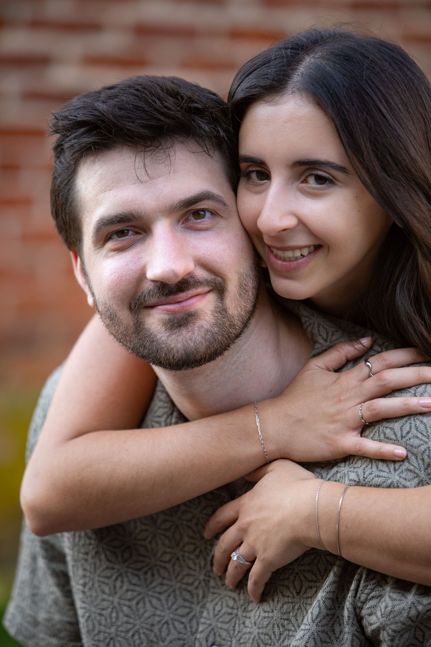 A happy couple embracing outdoors against a brick wall background, smiling warmly at the camera.