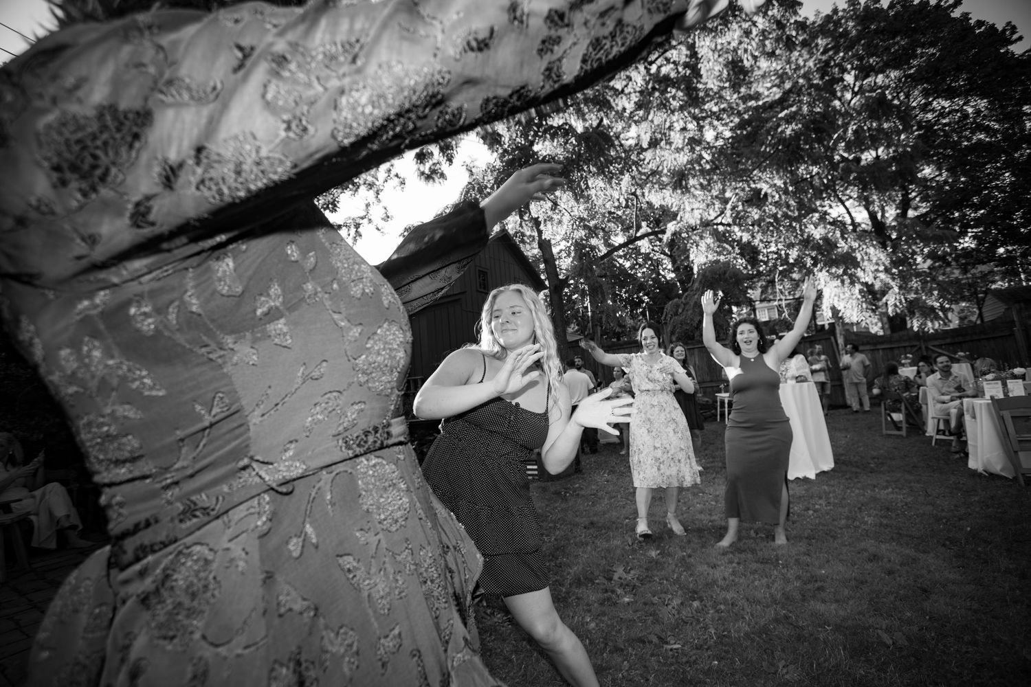 People dancing together in black and white at an outdoor celebration or wedding reception under trees.