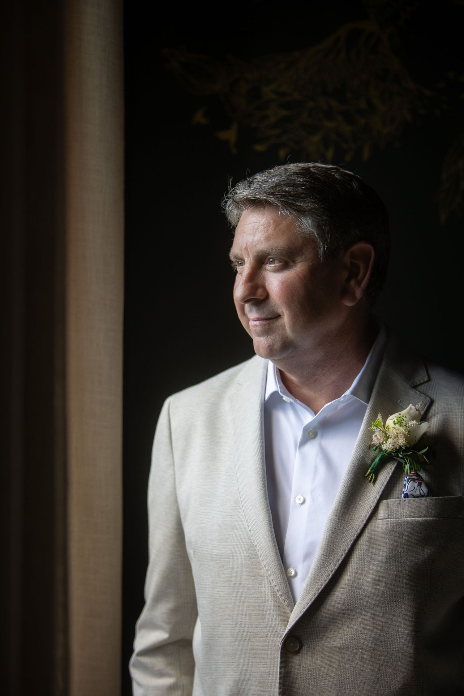 Groom in light suit and boutonniere poses by window in dramatic lighting.