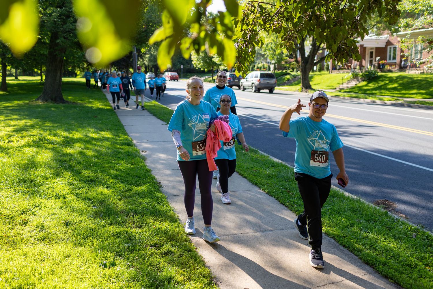 People walking along a tree-lined path during a community event on a sunny day.