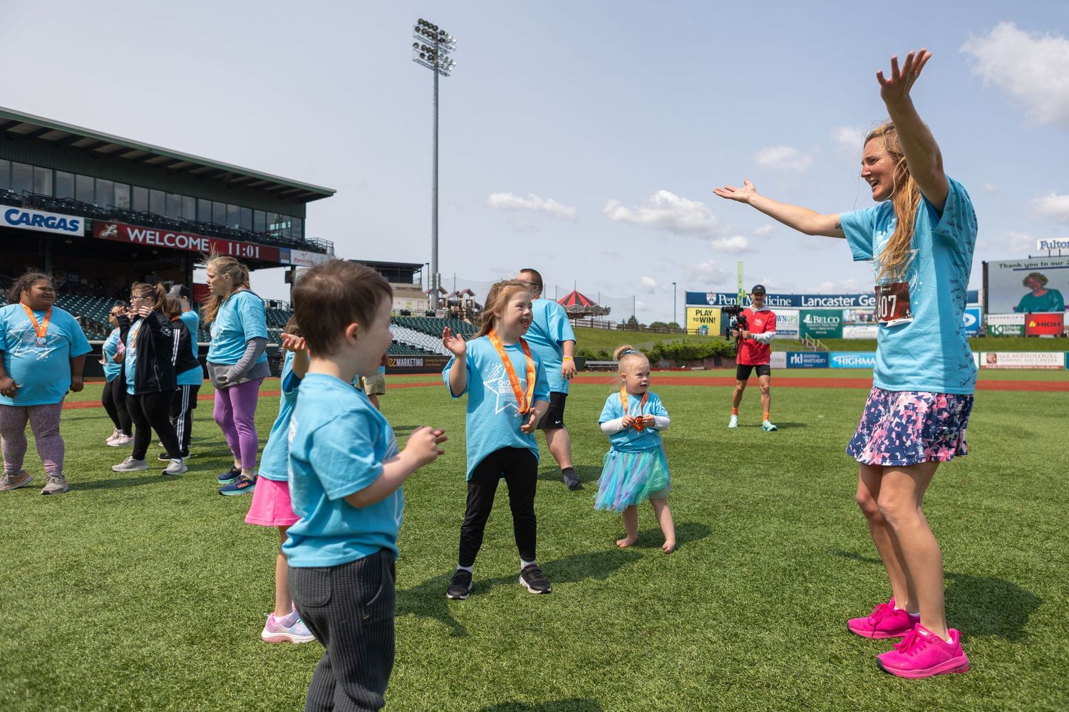 Children and adults participate in an activity on a sports field wearing matching blue shirts.