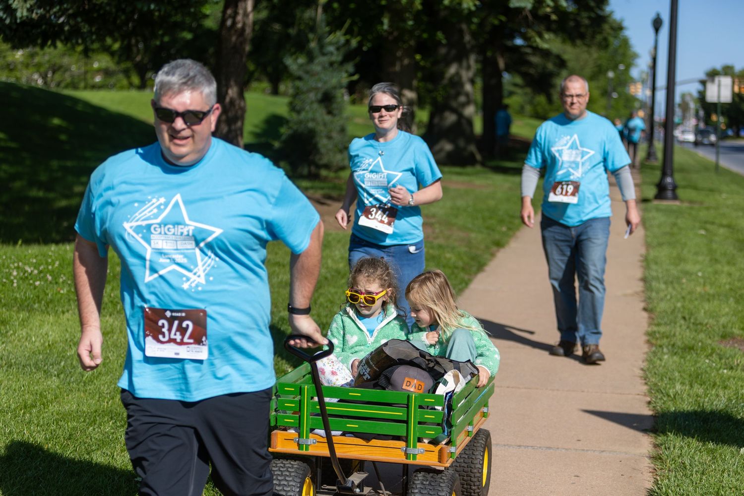 Participants in blue shirts walk along a path with a child riding in a small green wagon.