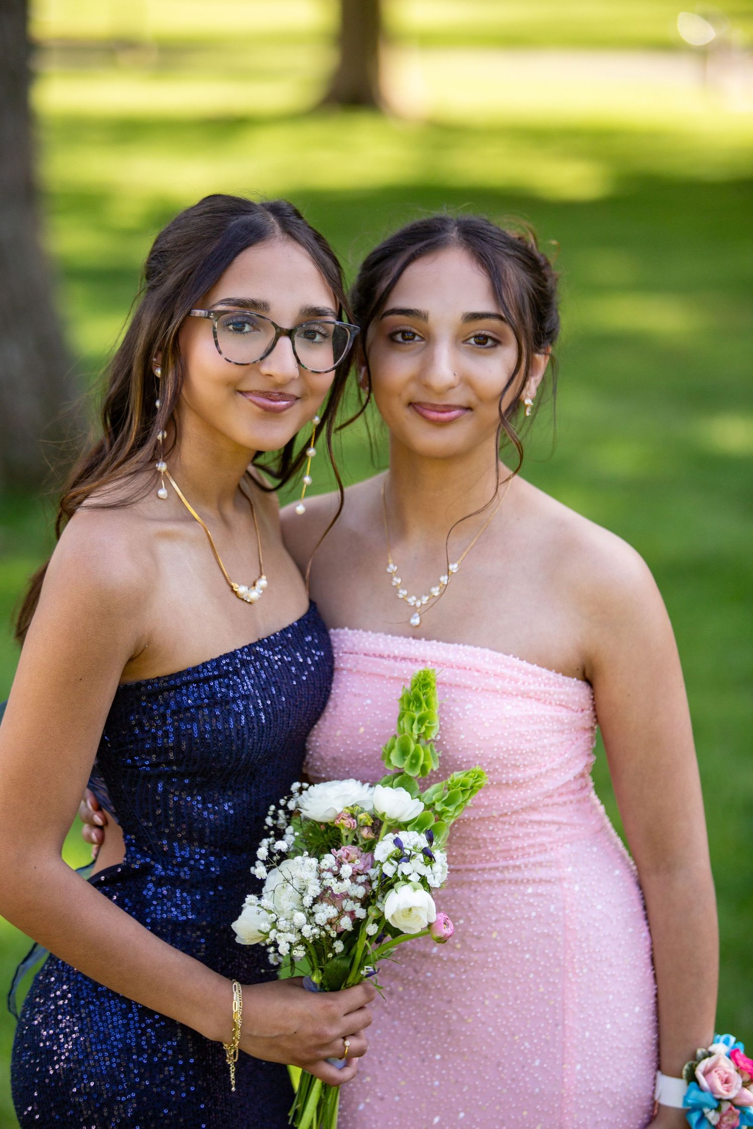Two women in formal dresses pose outdoors on a sunny day while holding a small floral bouquet.