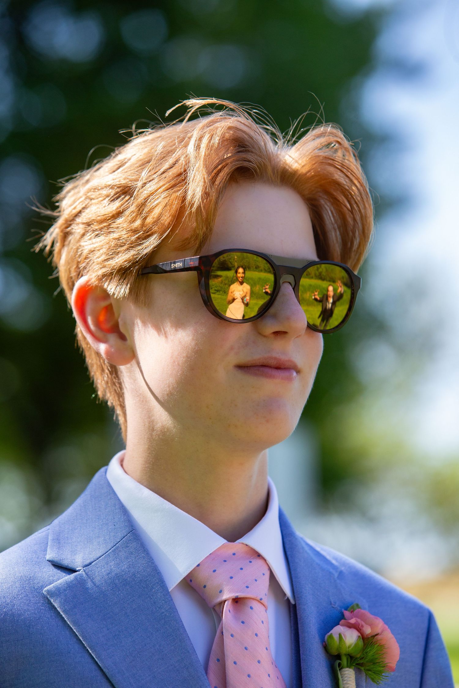 Someone wearing a blue suit with pink tie and reflective sunglasses stands outdoors in natural lighting with a boutonniere.