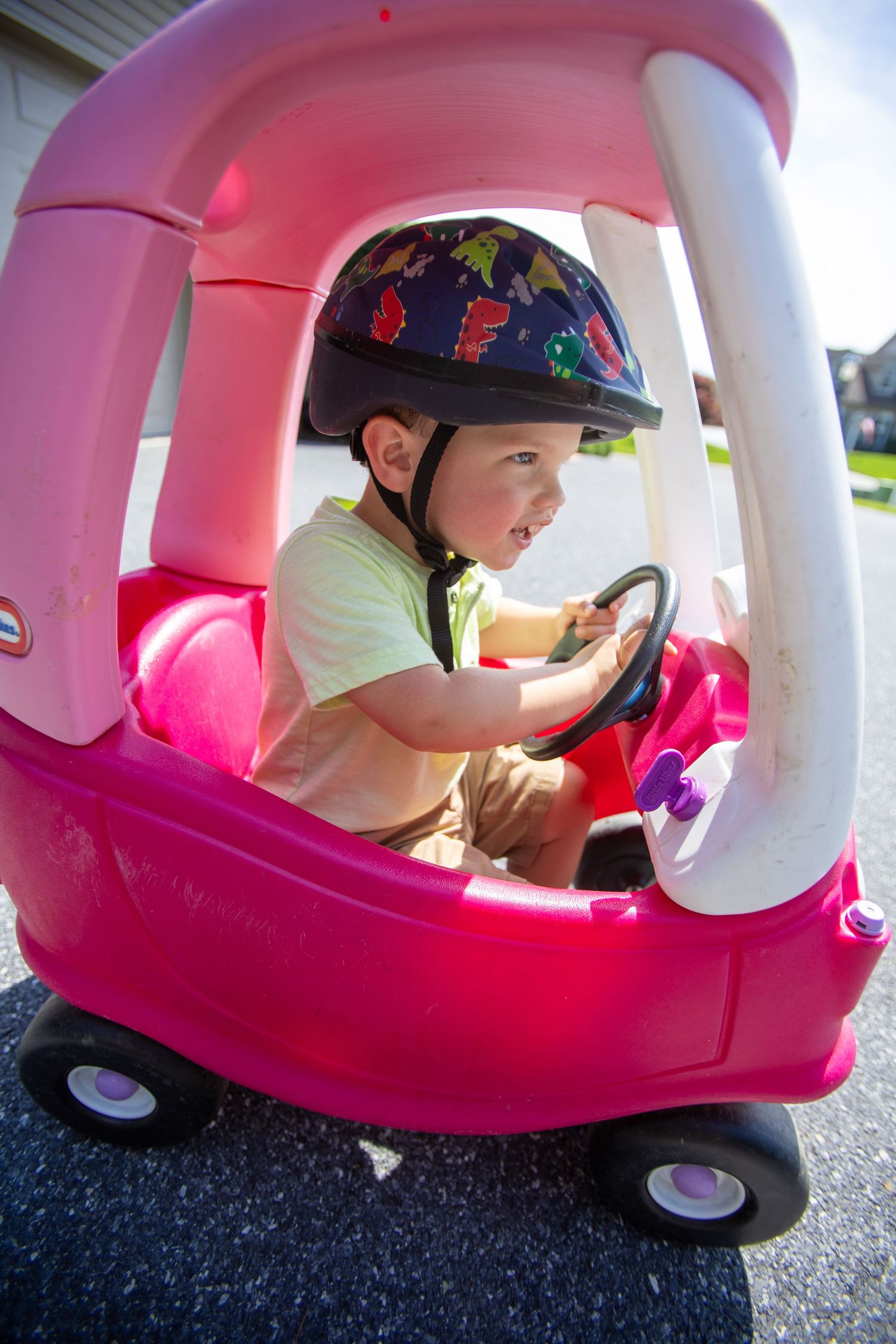 Child in a multicolored helmet plays inside a pink toy coupe car on a paved surface.