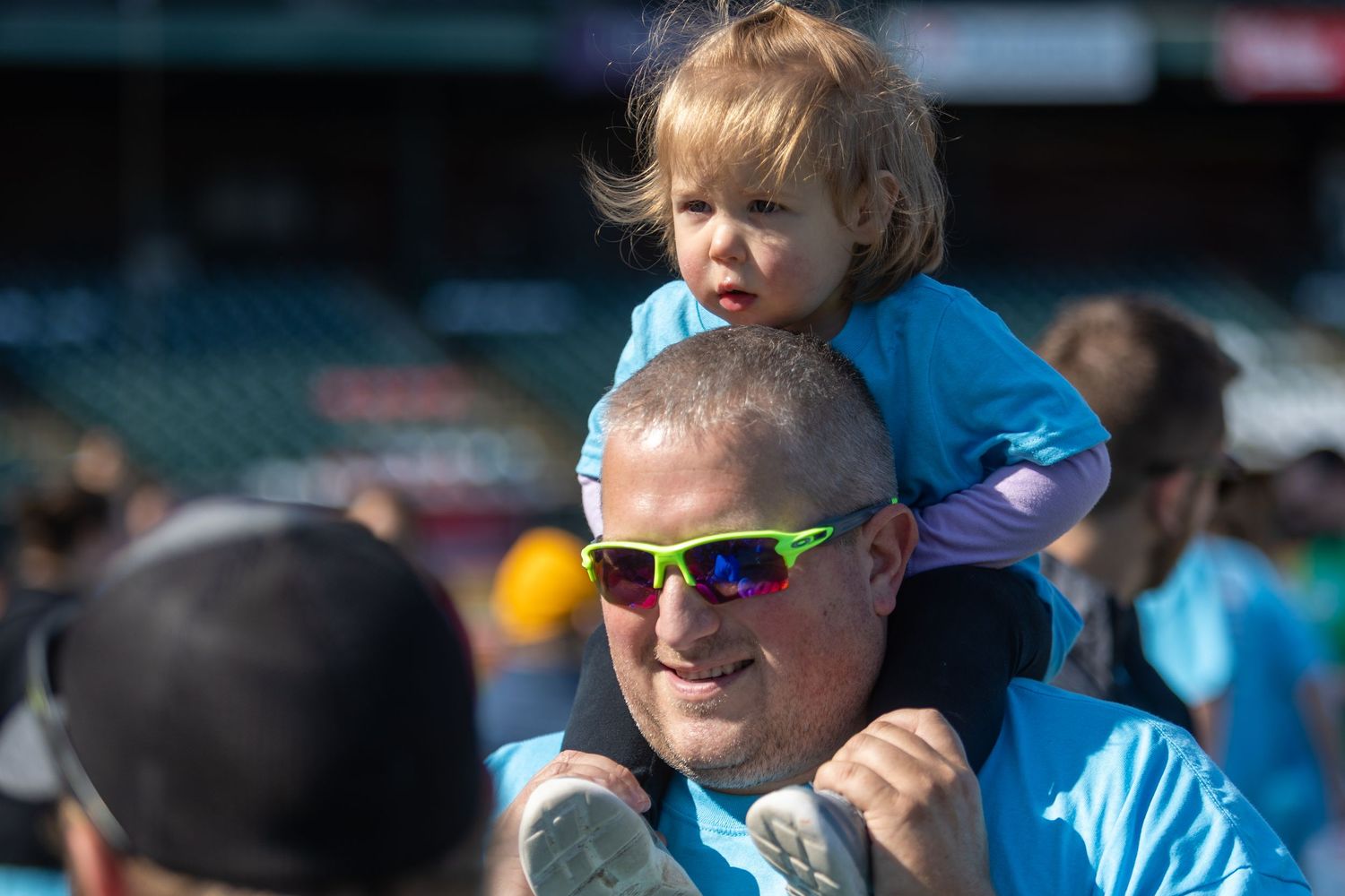 Person wearing sunglasses carrying child on shoulders at outdoor event.