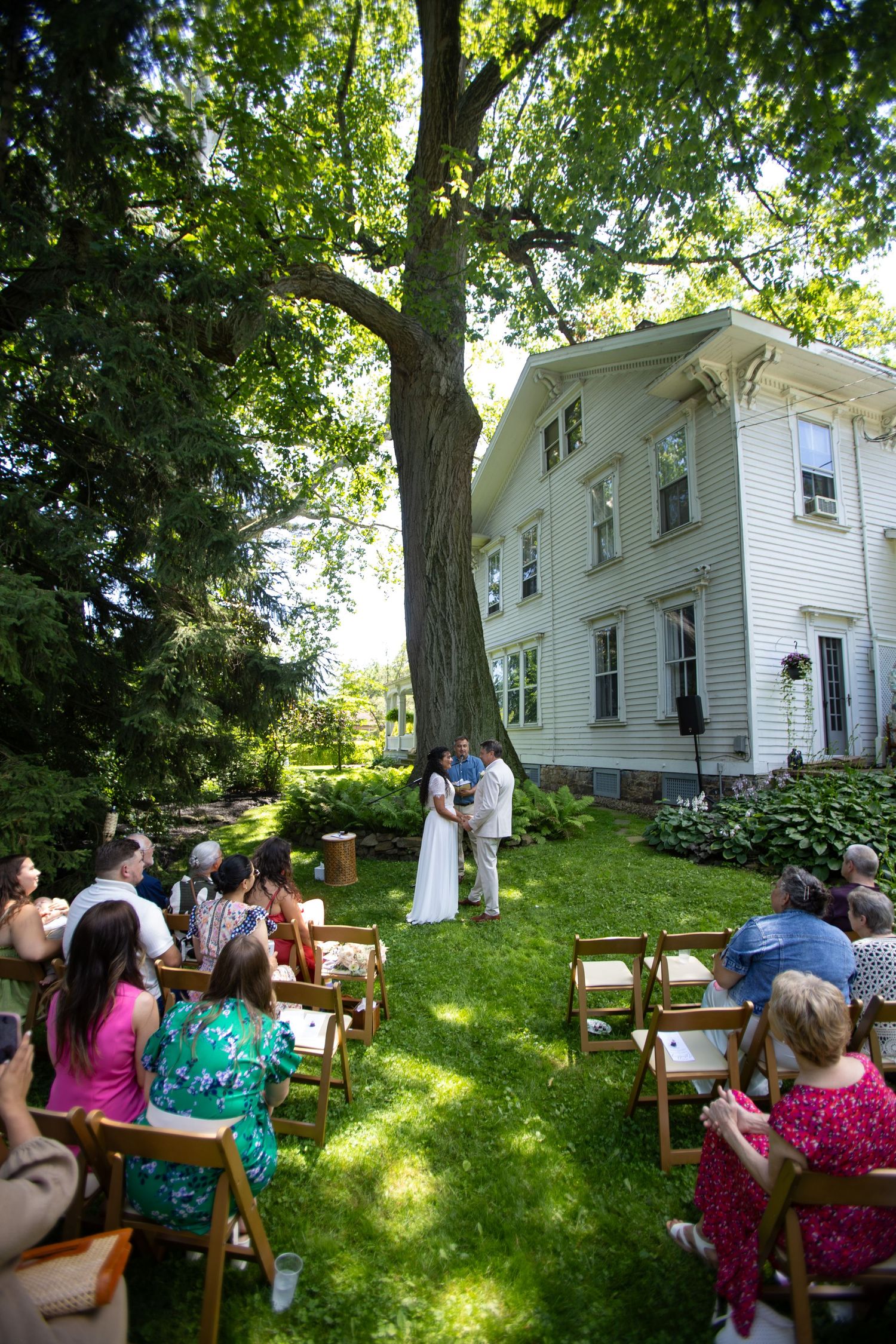 An intimate backyard wedding ceremony at a white farmhouse under a large tree with guests seated on wooden chairs.