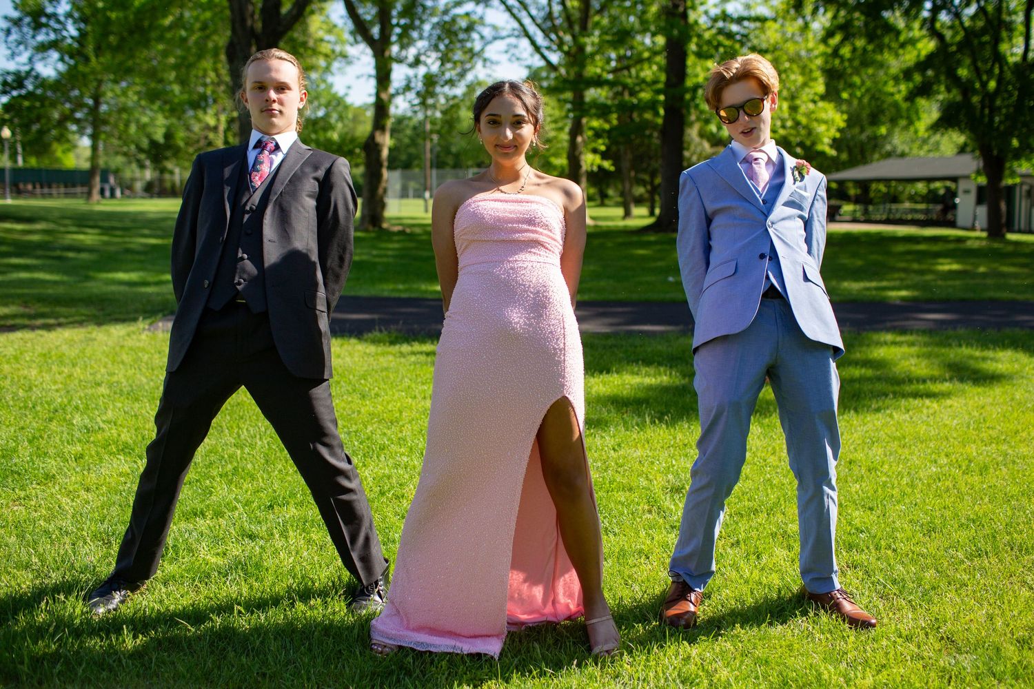 Three young people dressed formally pose together on a sunny lawn, with a person in a pink gown between two in suits.
