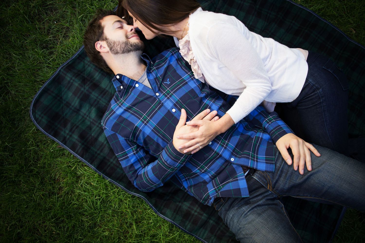 Couple lying together on a plaid blanket in green grass sharing an intimate moment.