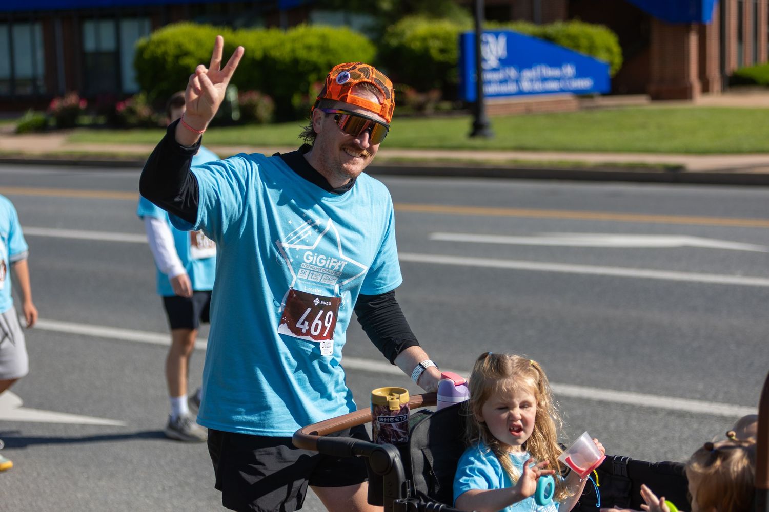 Participants wearing light blue shirts wave and smile during an outdoor community race event.