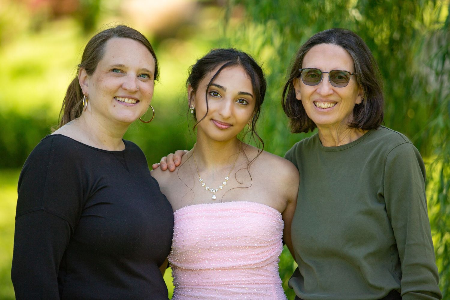 Three friends stand together outdoors on a sunny day, with one wearing a black top, one in pink, and one in olive green.