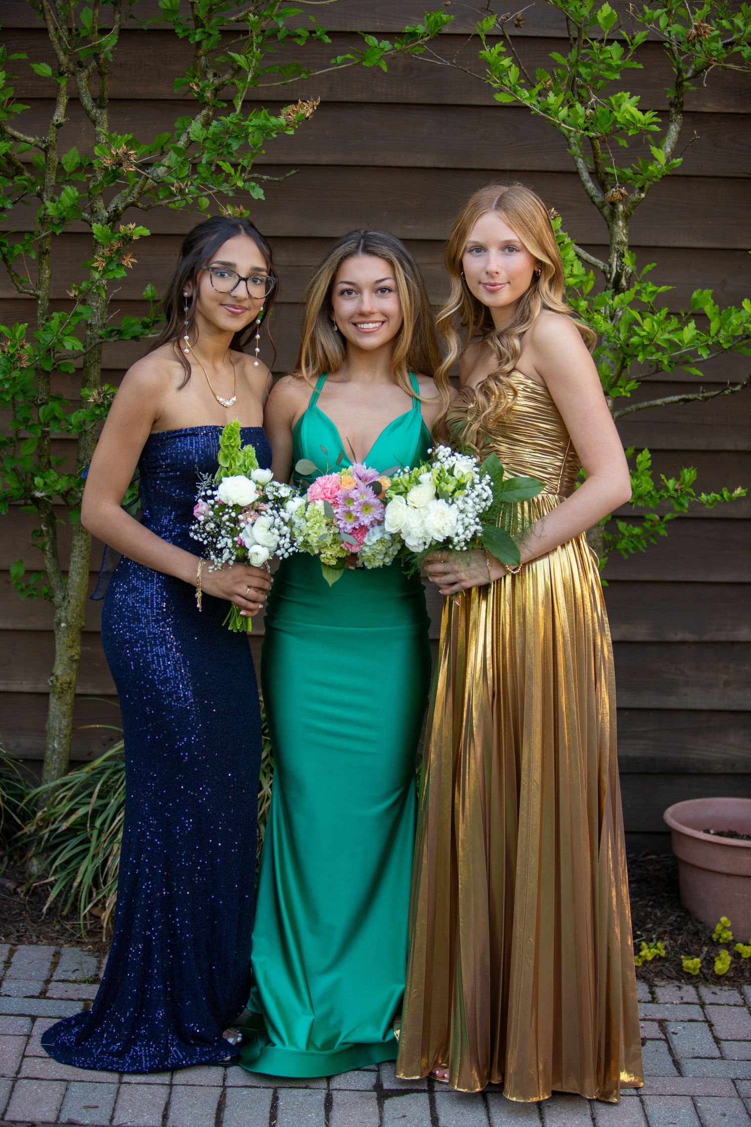Three friends in formal dresses - navy sequin, emerald green, and metallic gold - pose with floral bouquets outdoors.