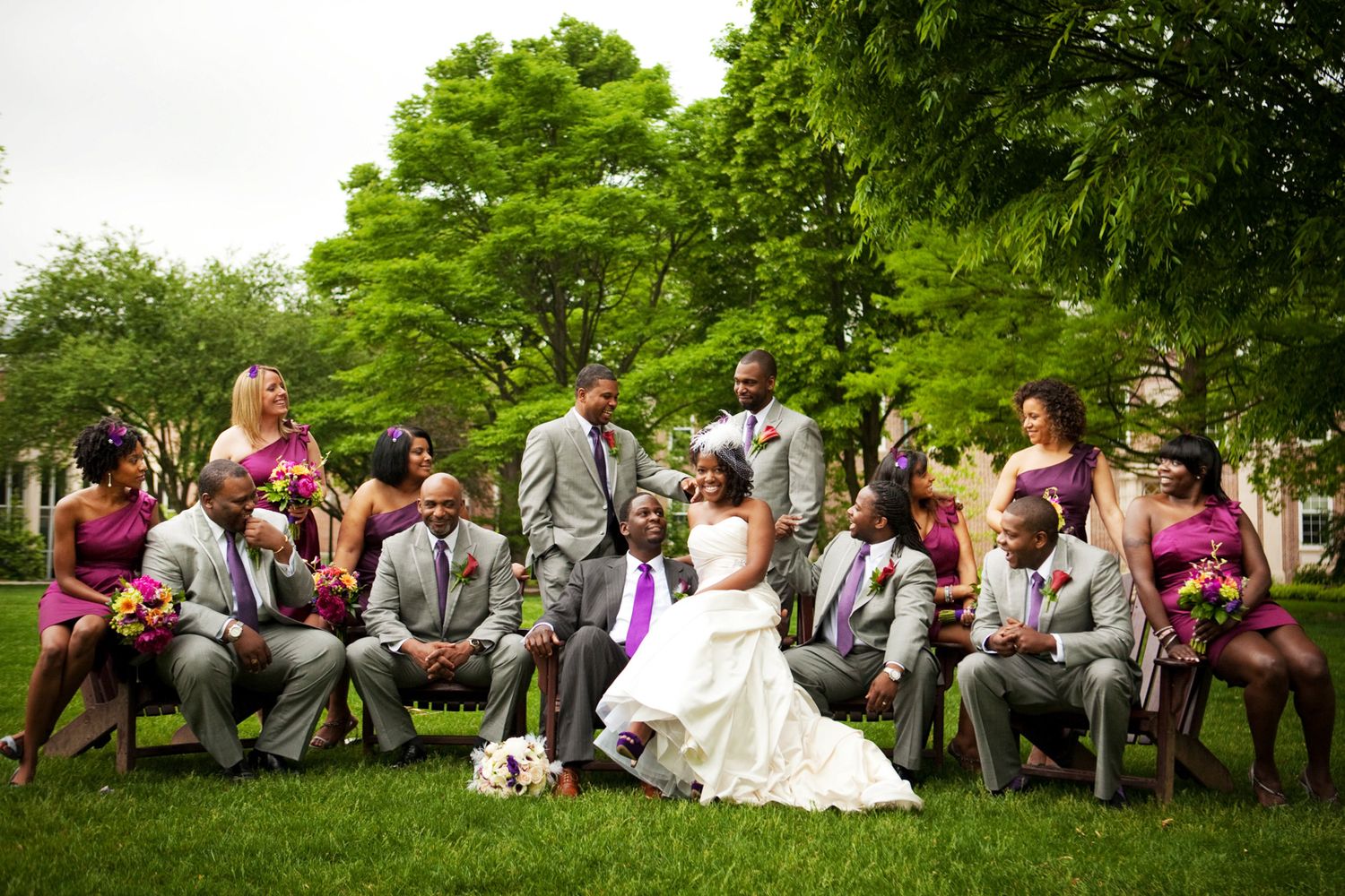 Wedding party lounges on chairs outdoors with bridal couple centered among trees and greenery.