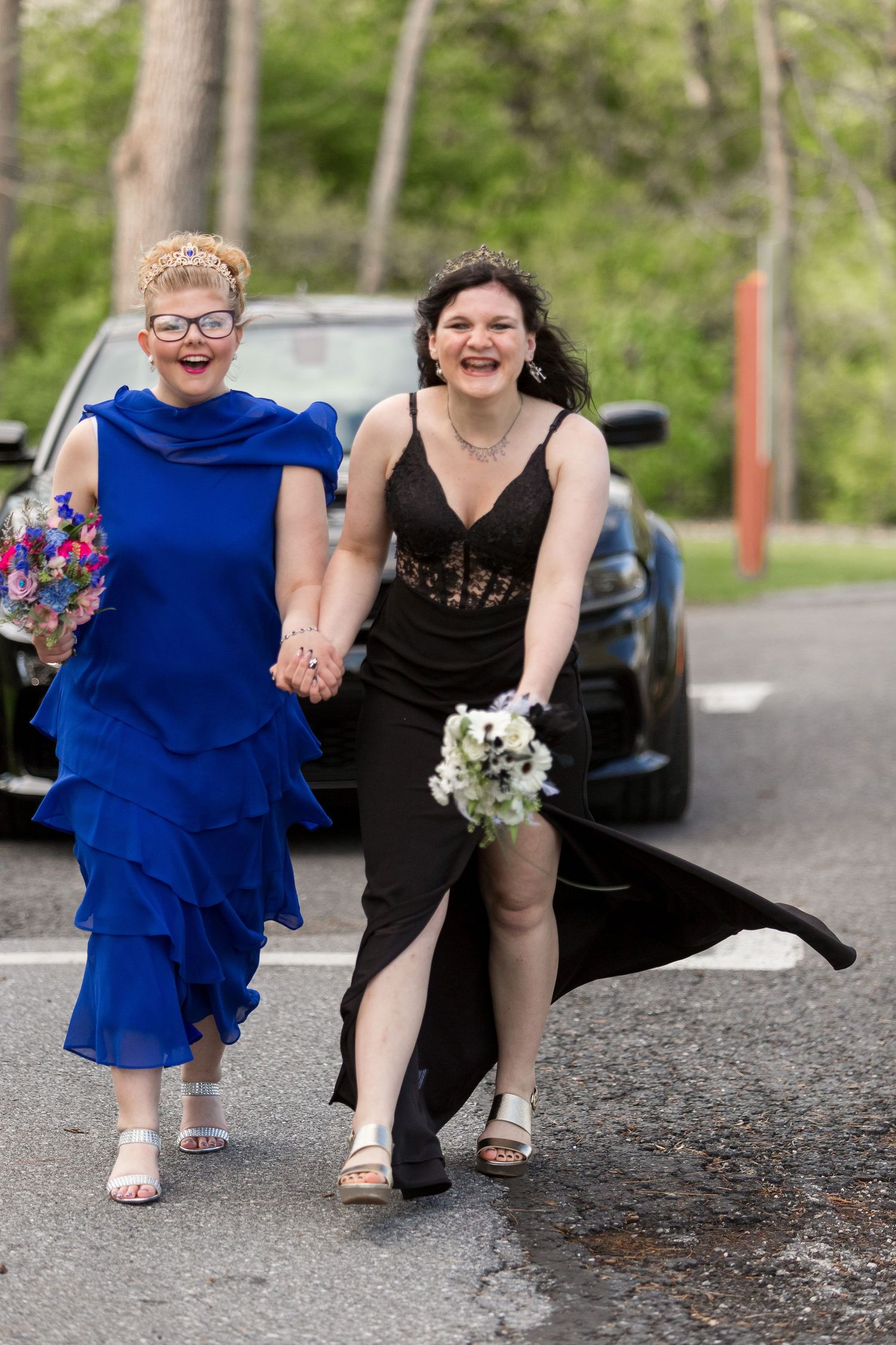 Two joyful prom attendees walk together, one wearing a ruffled blue dress and one in a black formal dress holding flowers, outdoors.