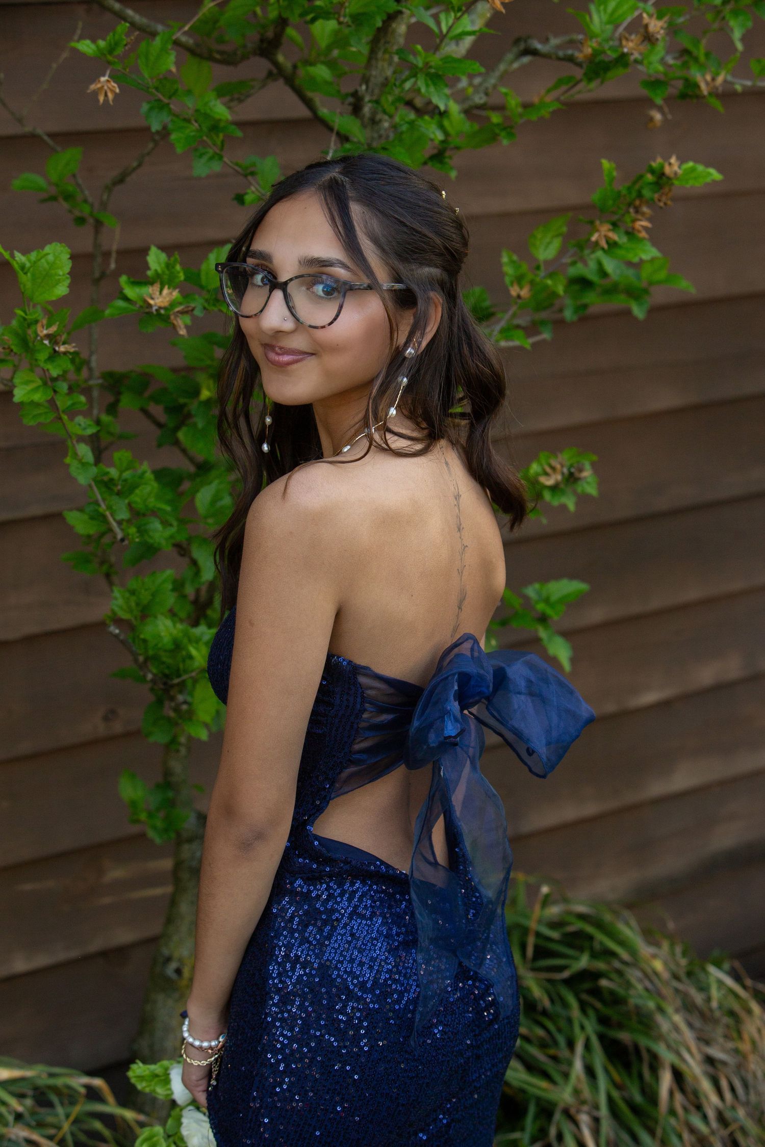 Elegant formal dress in sparkling navy blue with open back bow detail photographed against wooden wall with green foliage.