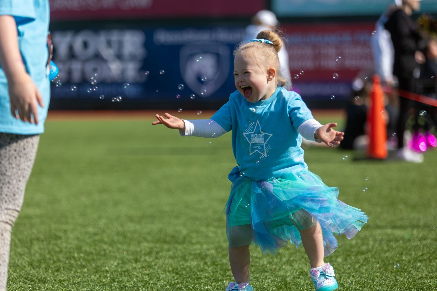 A young child in a light blue shirt and tutu twirls joyfully on a sports field.