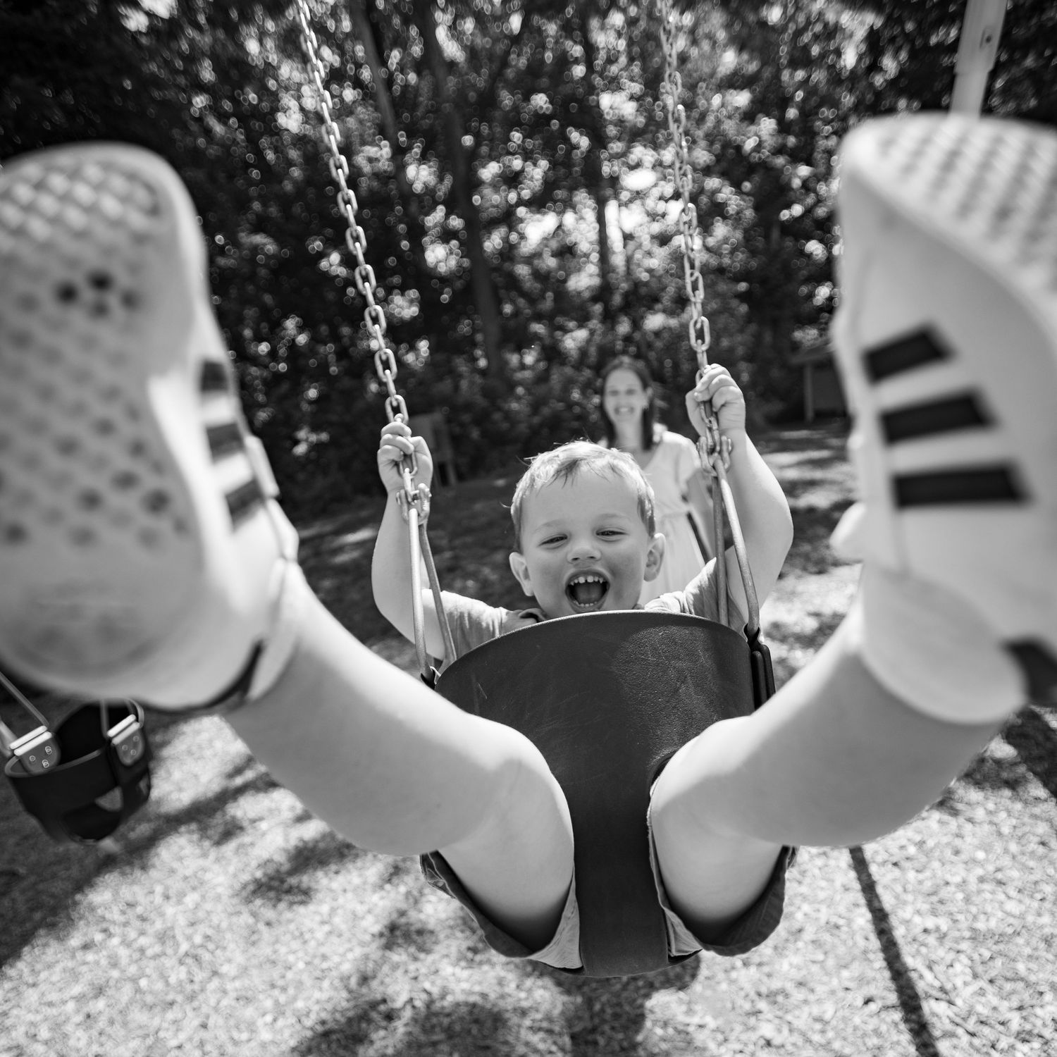 A joyful child swings at a playground with feet in white sneakers pointed at the camera in black and white photography.