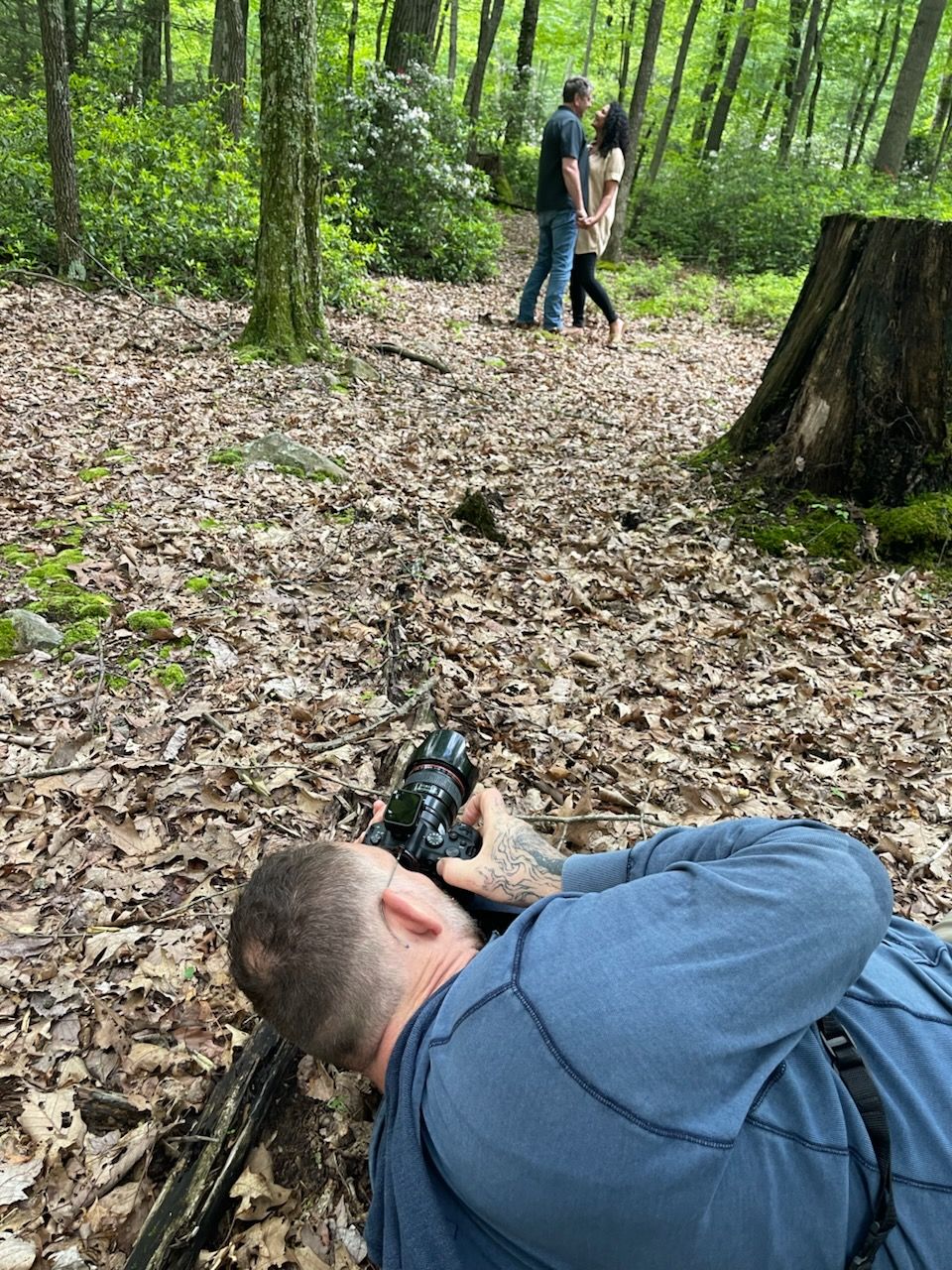 A photographer lies prone on leaf-covered forest ground while taking pictures of a couple on a wooded trail.