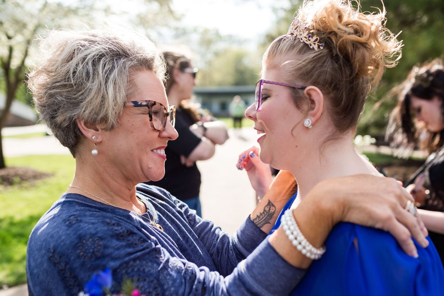 Two people share an emotional moment outdoors, embracing while one wipes tears in bright sunlight.