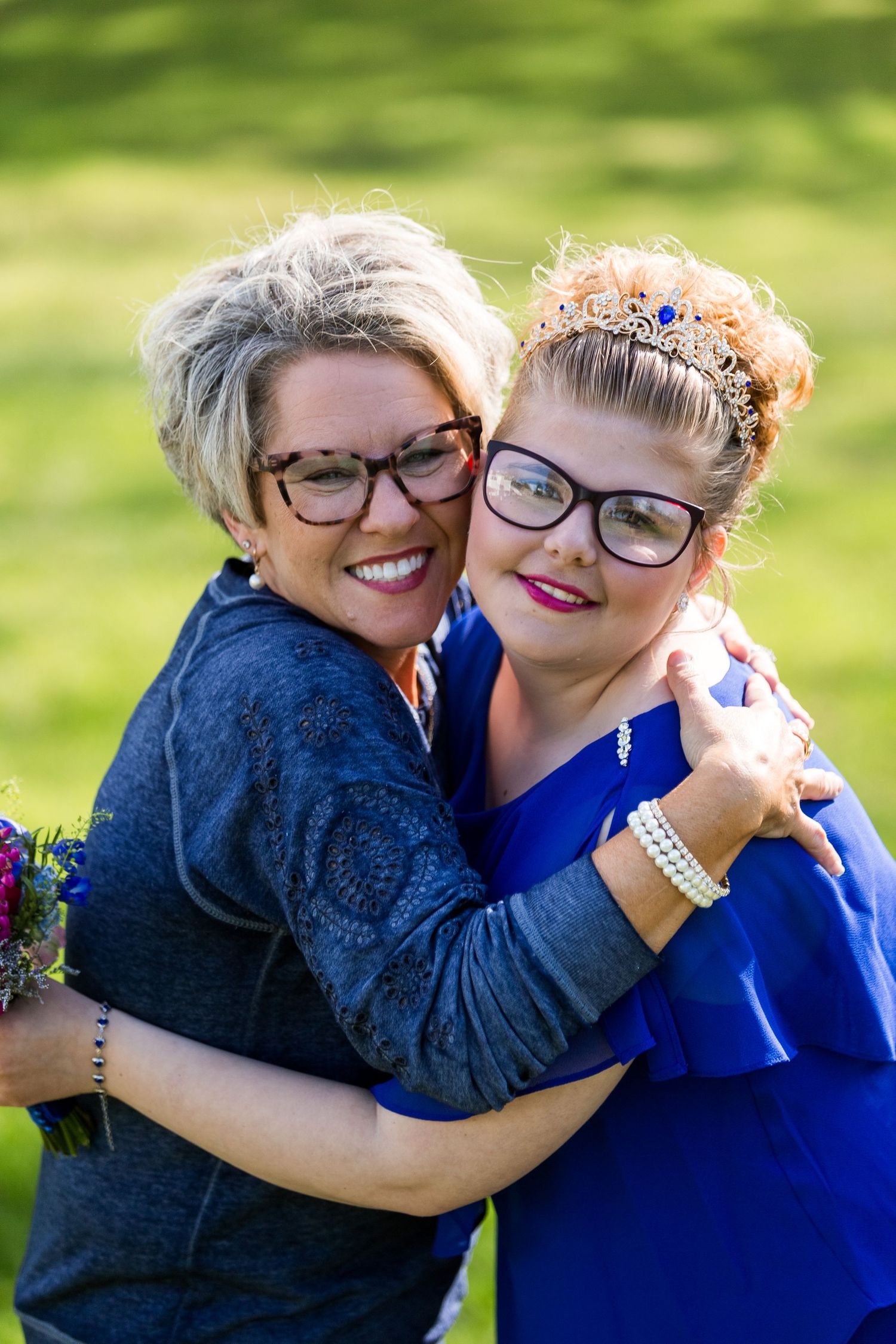 Two people wearing glasses embrace and smile outdoors, one in blue dress and one in dark top on sunny day.