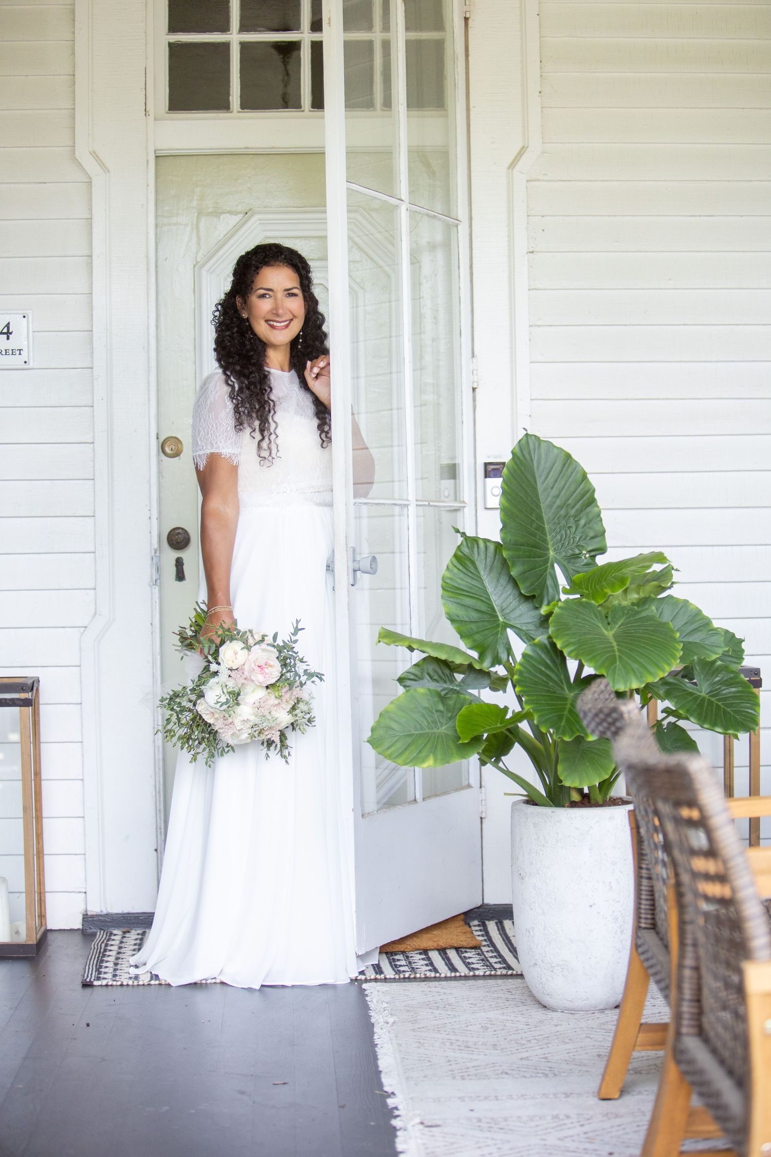 A bride in a white wedding dress poses with her bouquet on a white porch with large tropical plants.