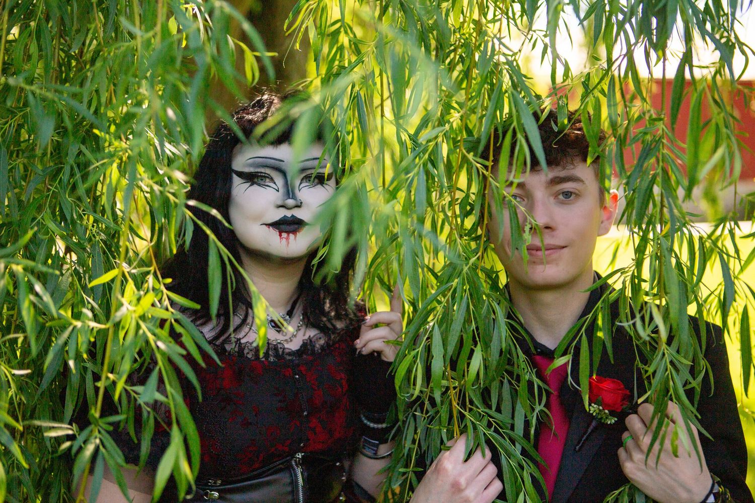 Two people with gothic style makeup and clothing pose among drooping willow tree branches in nature.