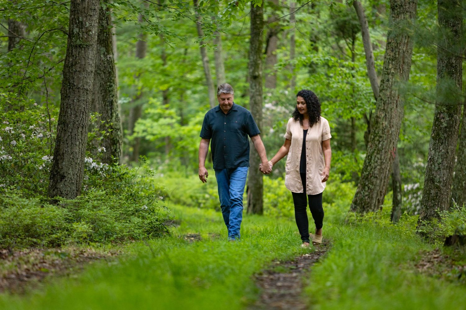 Two people walk hand in hand along a lush green forest trail surrounded by tall trees and verdant foliage.