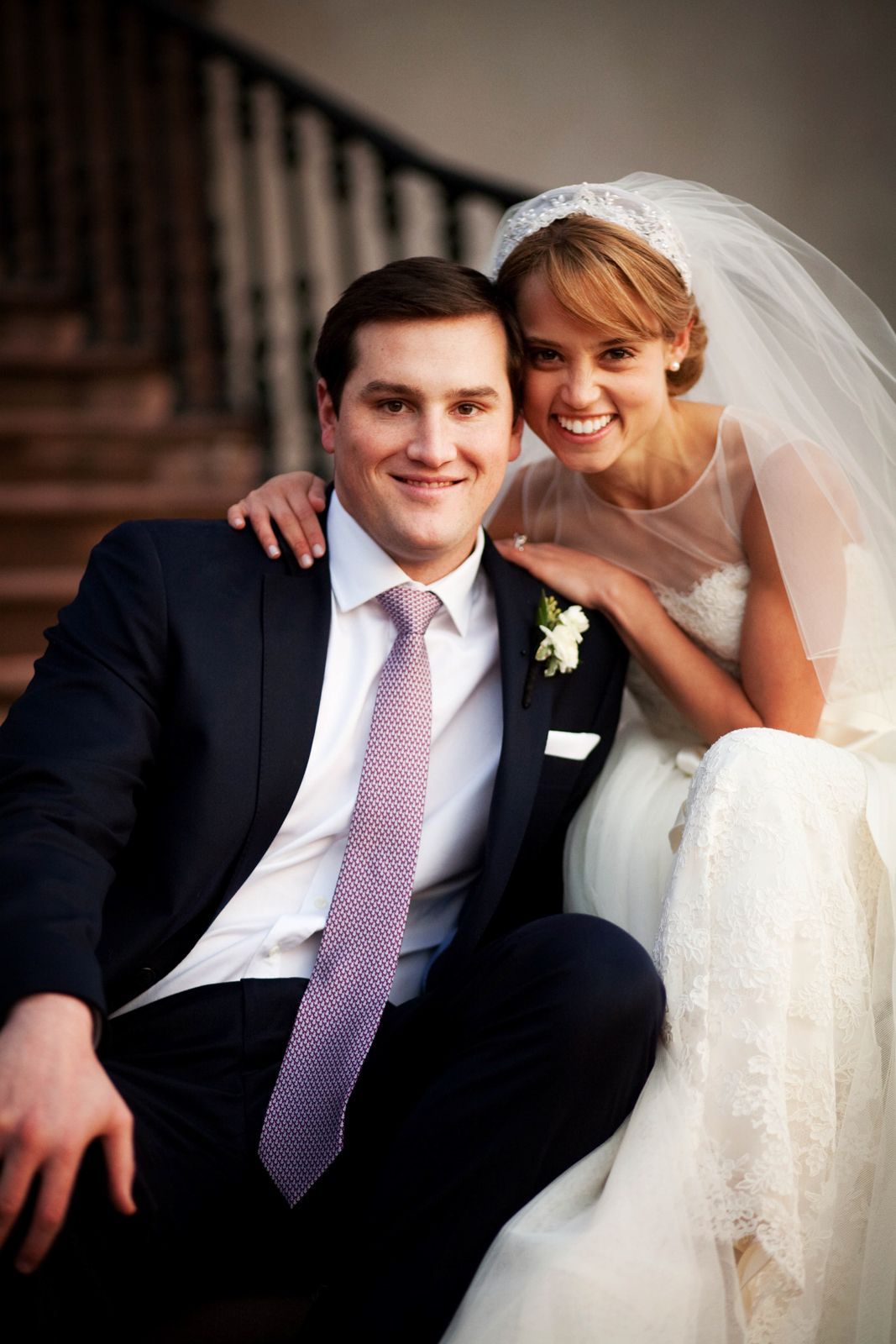 Wedding couple in formal attire sitting on stairs, with bride in white dress and veil and groom wearing navy suit and pink tie.