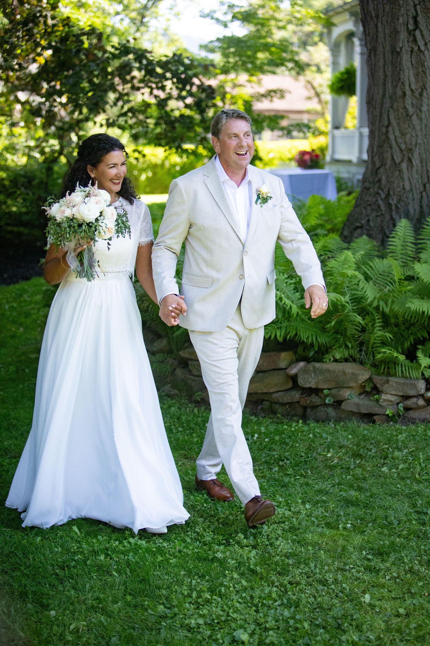 A wedding couple in all white attire stroll joyfully through a lush garden with ferns and greenery.