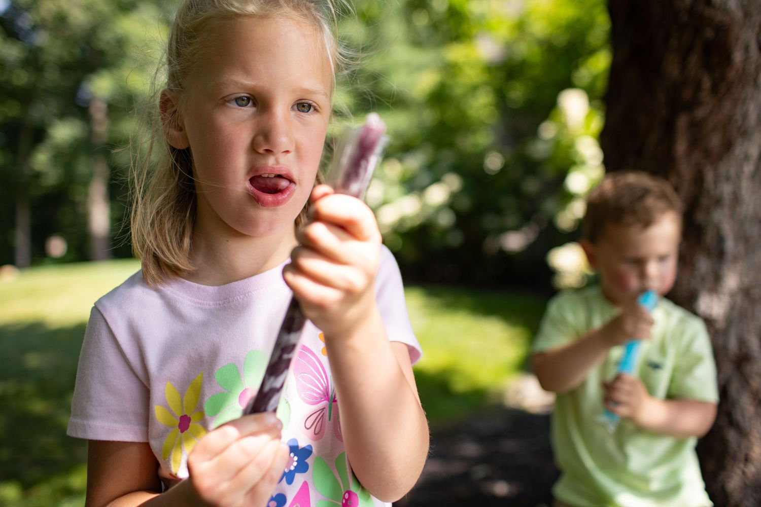 Children enjoy ice cream cones while standing outdoors in a sunny garden setting.