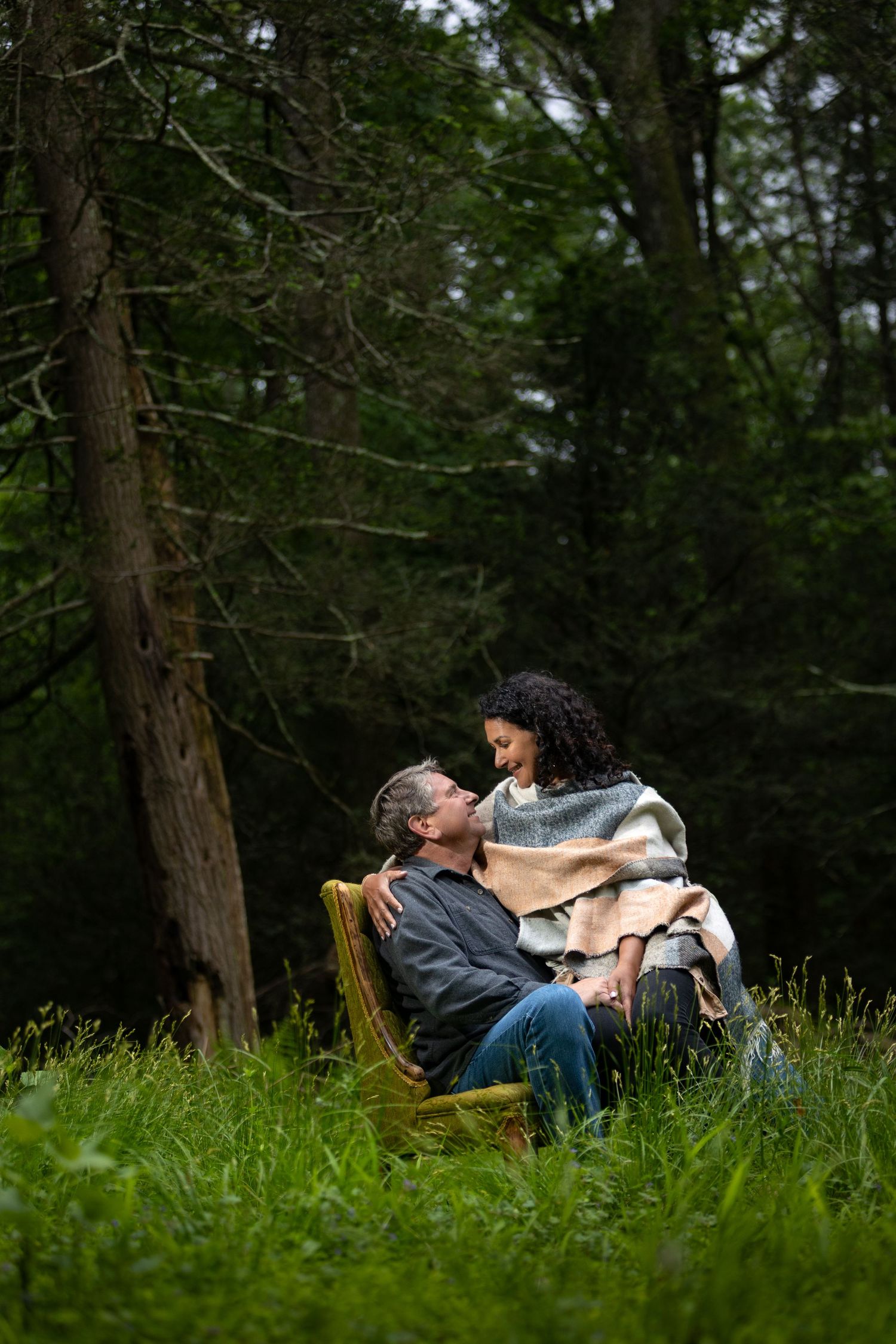 A couple shares an intimate moment while sitting on a wooden chair in a lush forest setting.