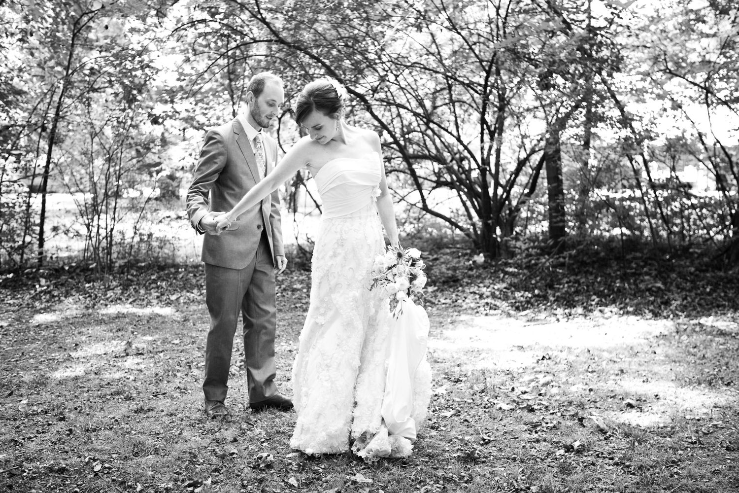 A couple in wedding attire walk together on grass with trees in the background in a black and white outdoor photograph.