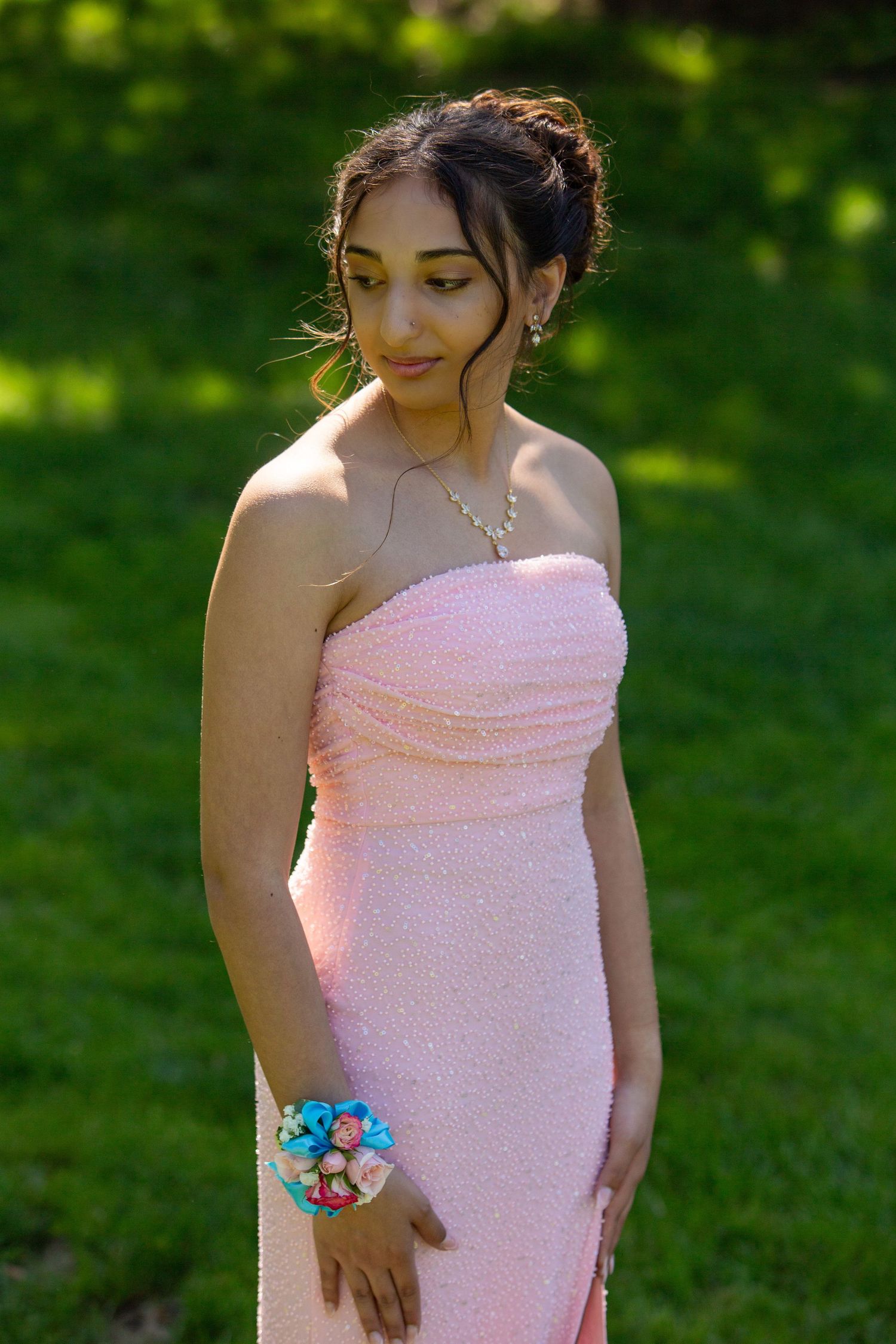 A person in a strapless pink prom dress poses outdoors on a sunny day wearing a blue floral wrist corsage.