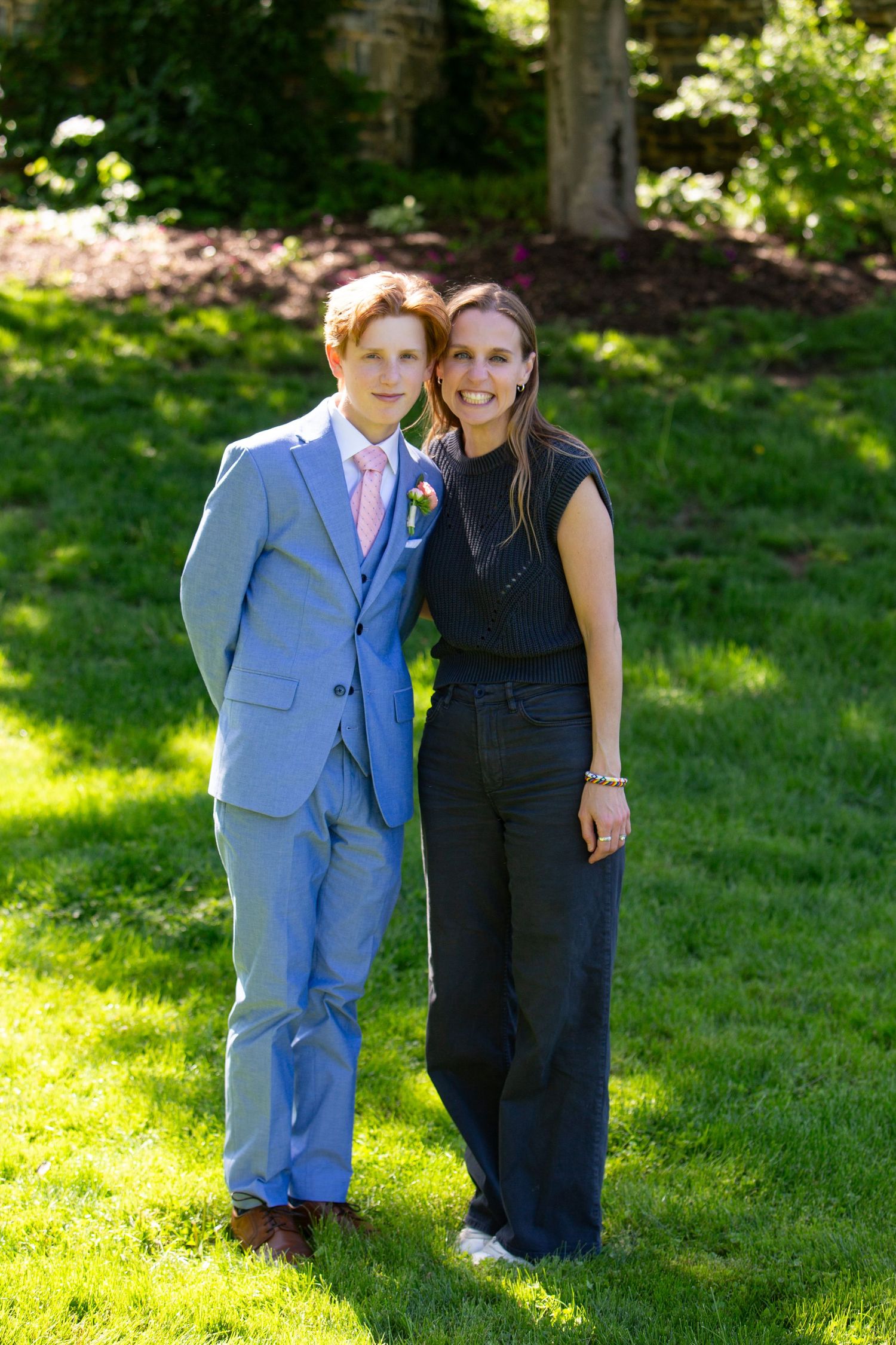 A couple poses together on a sunny lawn, one wearing a light blue suit with pink tie and one in a navy jumpsuit.