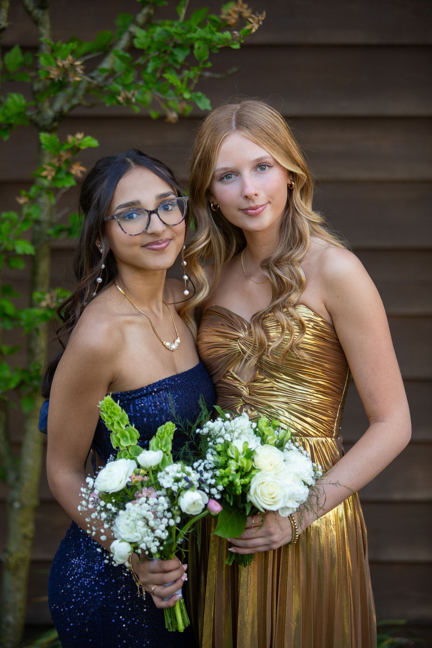 Two friends in formal dresses - one in navy sequins and one in gold - pose together holding white rose bouquets.