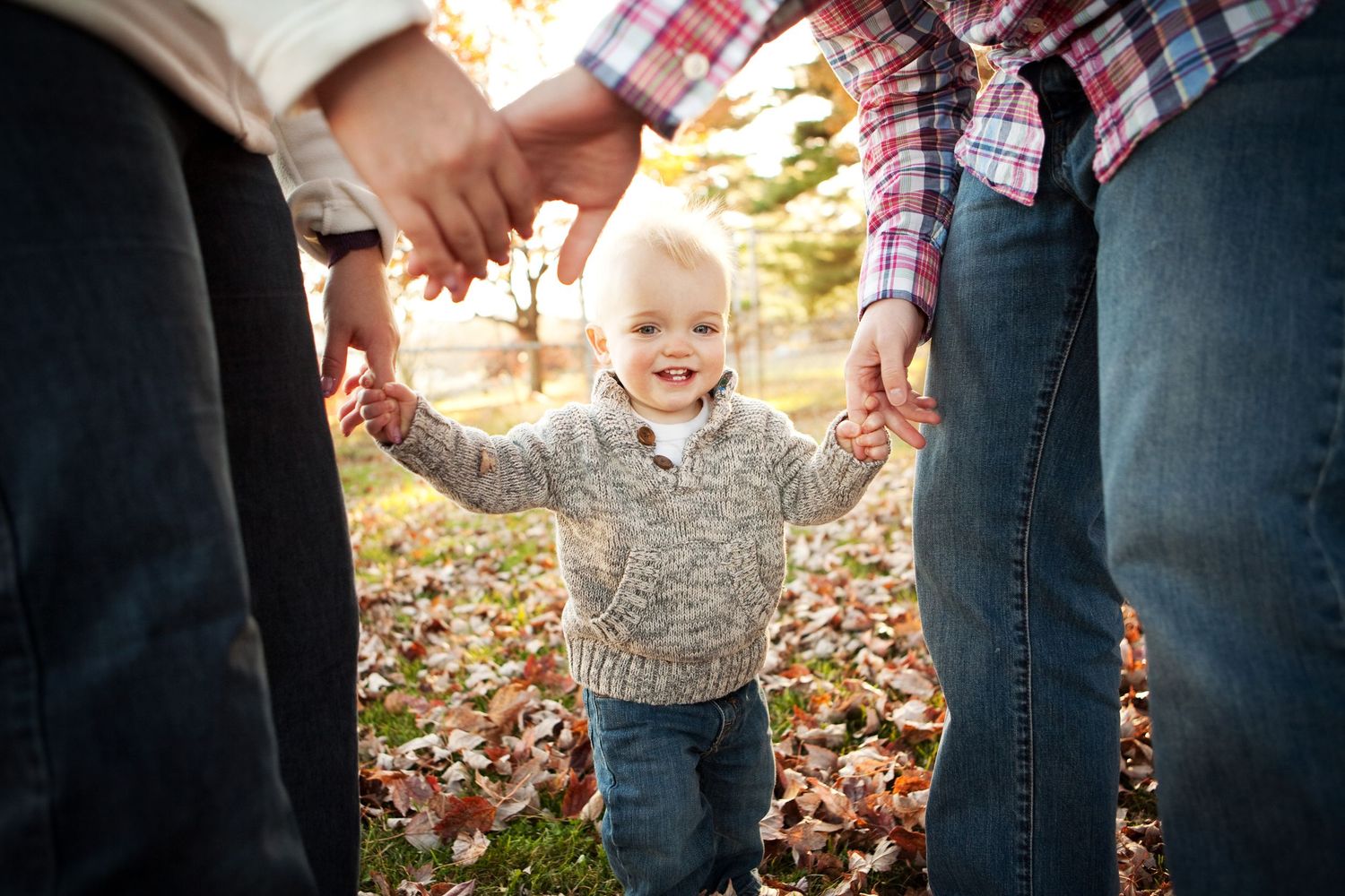 A joyful child in a gray sweater walks while holding hands with parents among scattered fall leaves on the ground.