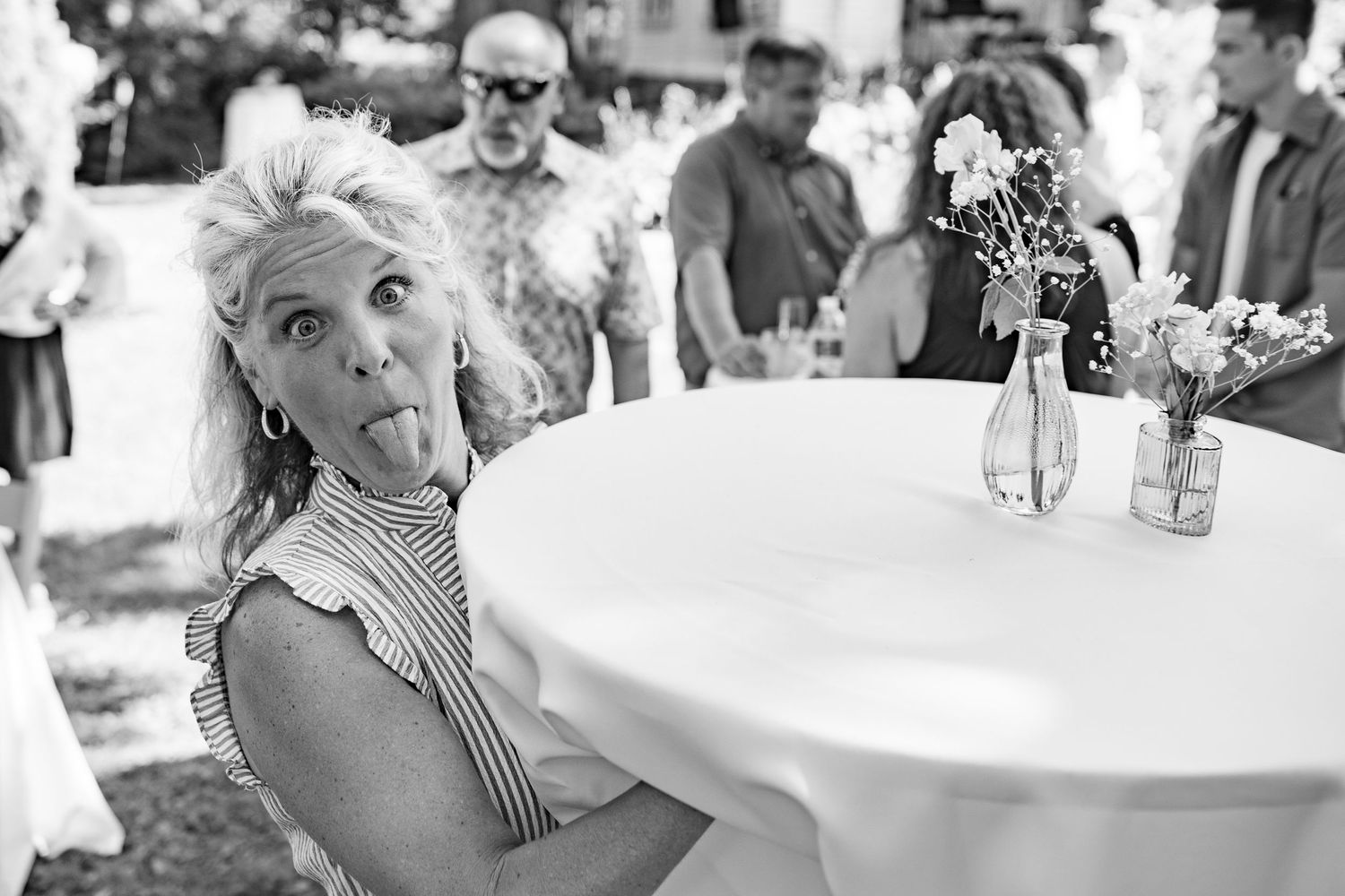 Black and white candid photograph of someone making a silly face at an outdoor party with a white round table.