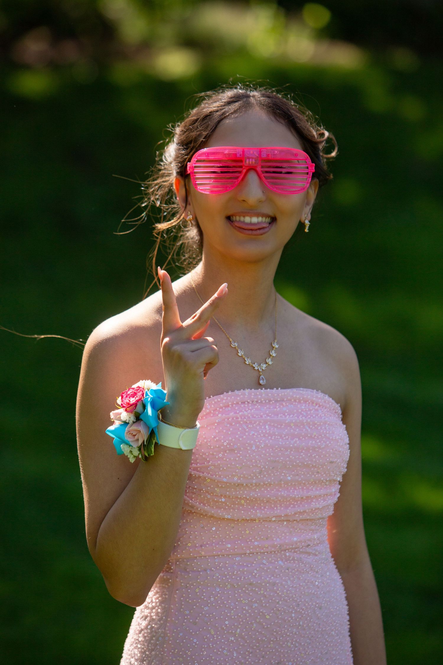 Person in pink strapless dress wearing hot pink shutter shades and colorful corsage makes peace sign outdoors.