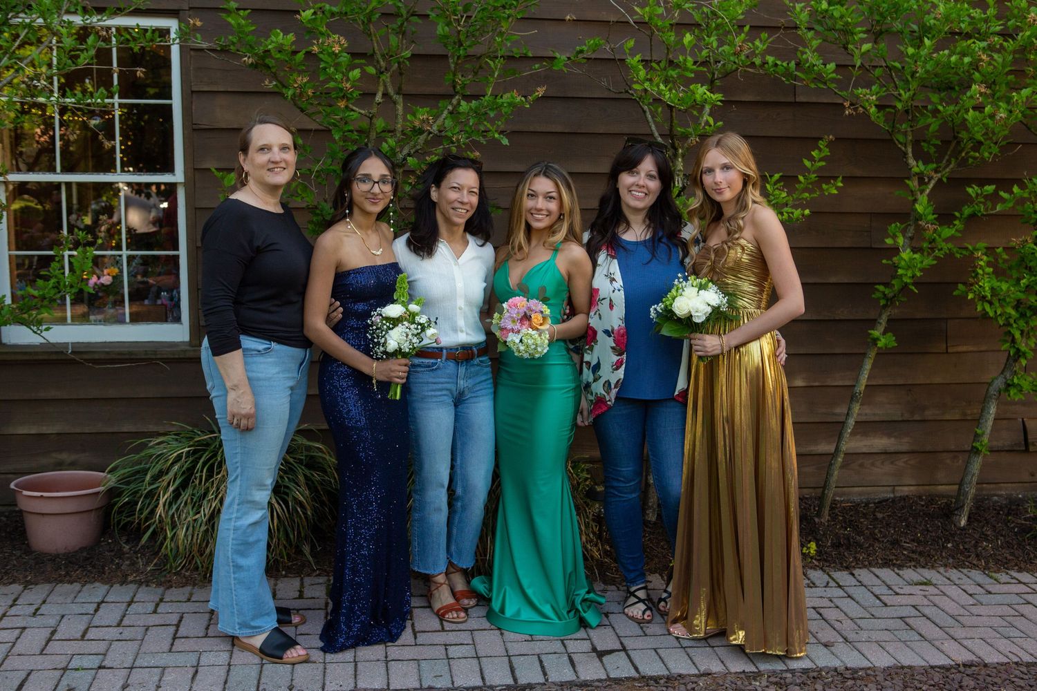 Group in coordinated outfits posing for a casual outdoor photo while holding bouquets against a wooden wall.