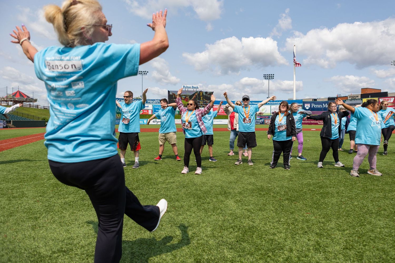 Group of participants in light blue shirts exercising together on a baseball field during an outdoor event.